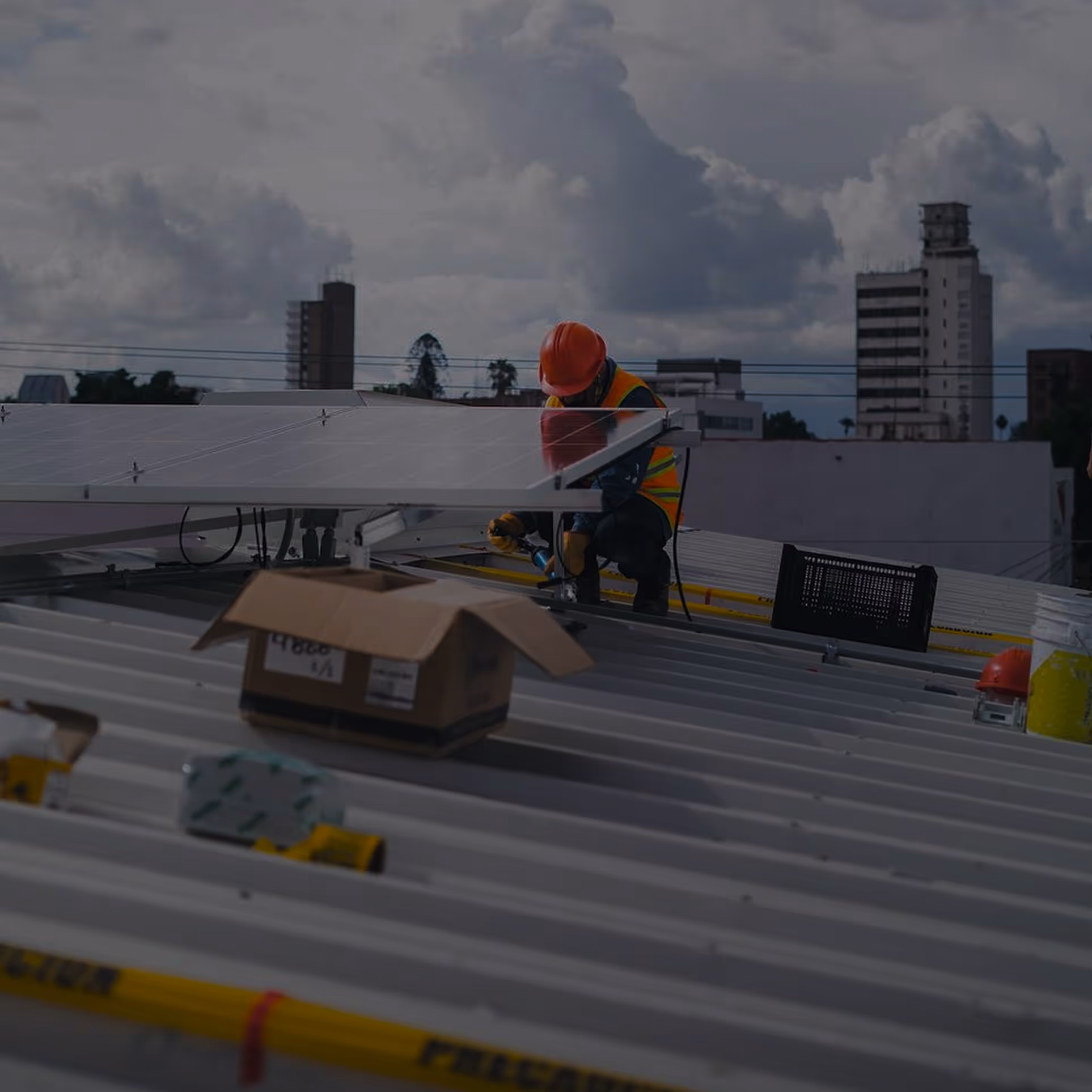 Worker wearing orange helmet and safety vest installing solar panels on a metal rooftop with city buildings and cloudy sky in the background.
