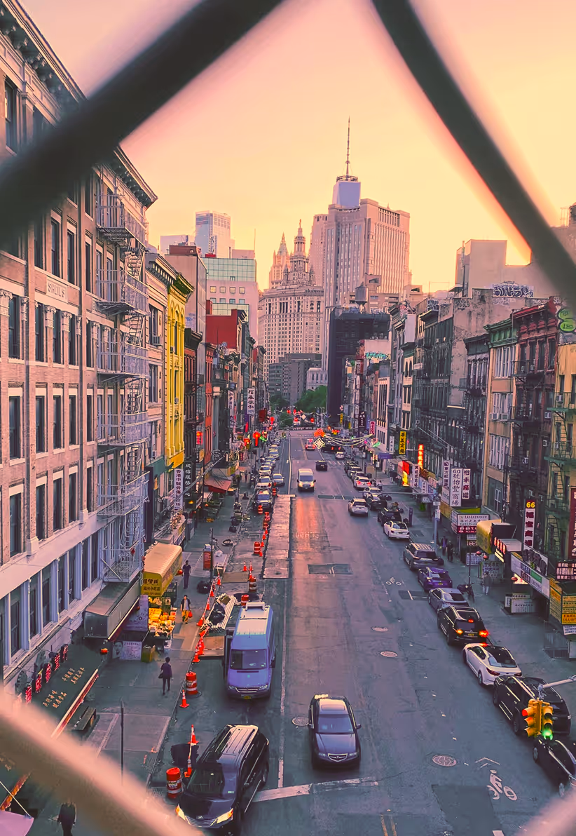 Street view of a busy city road lined with buildings and cars, framed by a blurred fence, with skyscrapers in the background at sunset.