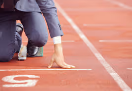 Person in business suit crouching at starting line on a running track marked with number 5.