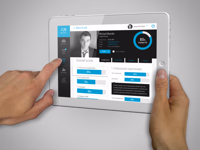 Person holding a white tablet displaying a job candidate profile with a photo, scores on professional requirements, personality traits, languages, and computer skills, showing an 85% perfect fit score.