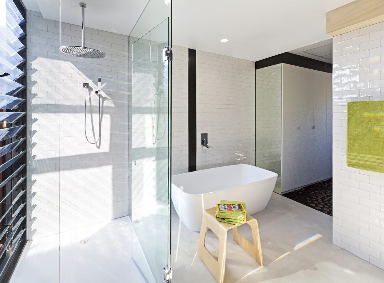 Bright modern bathroom with a glass shower enclosure, white freestanding bathtub, wooden stool with green towels and a book, and white tiled walls.