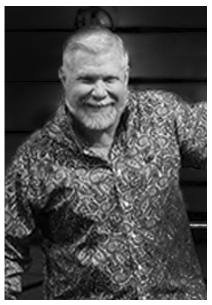 Smiling man with short hair and beard wearing a patterned shirt standing in front of a dark background.