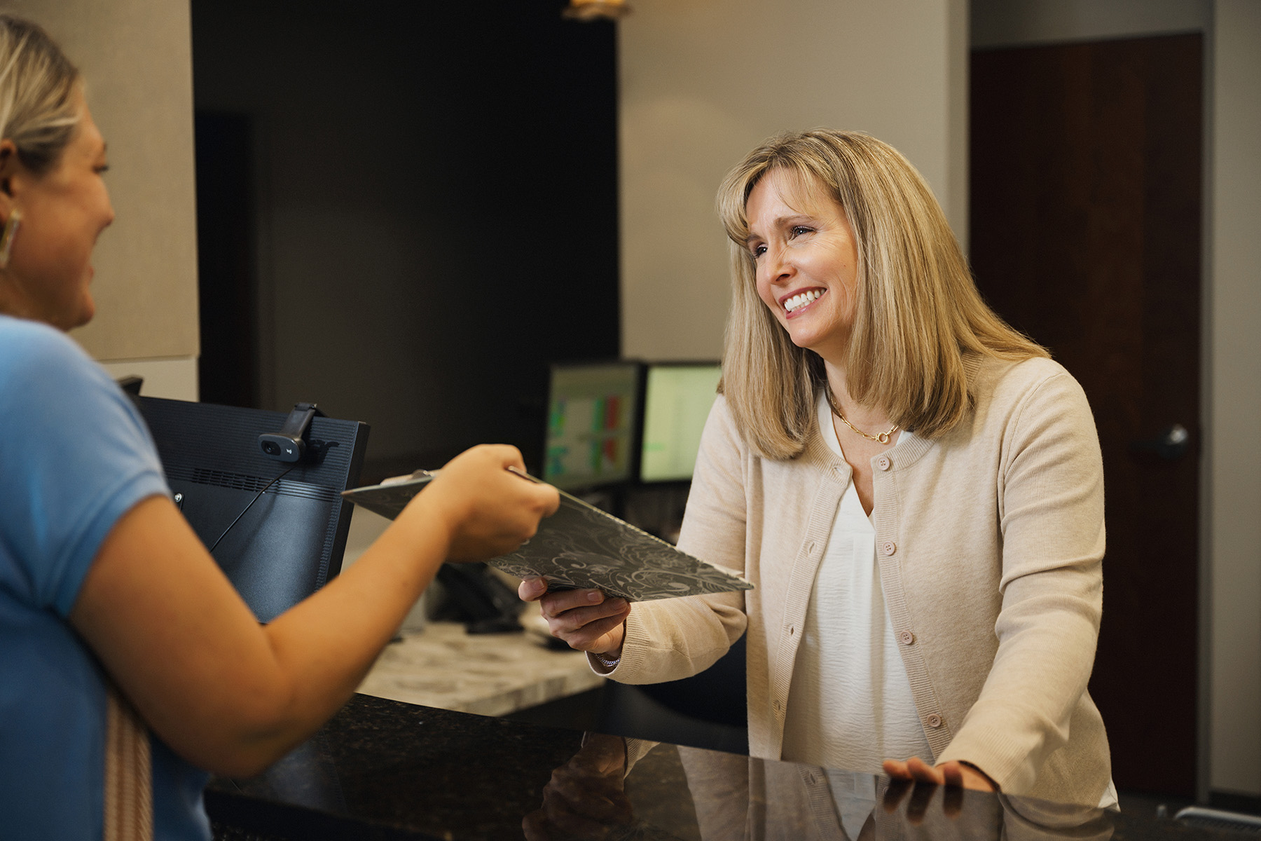 Smiling receptionist handing a clipboard to a woman at an office front desk.