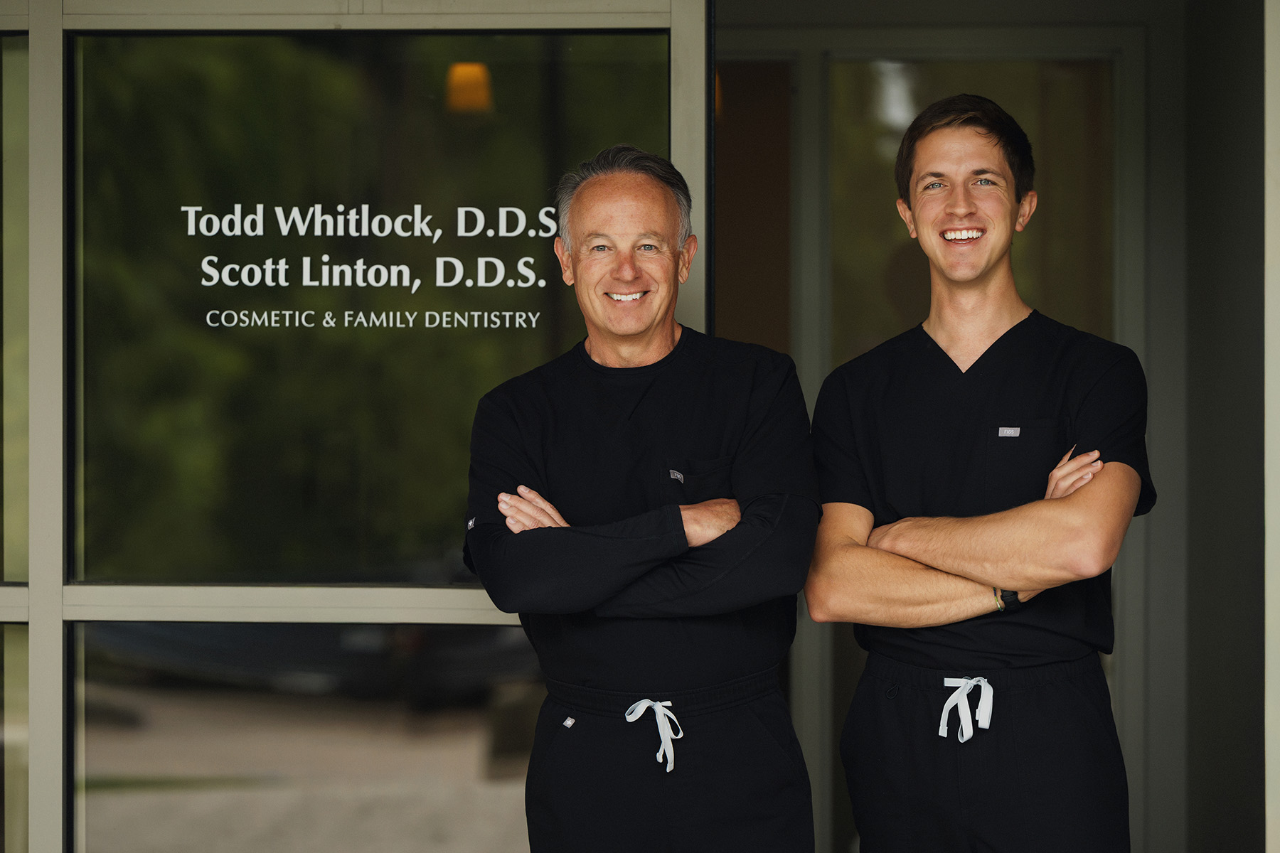 Two male dentists in black scrubs standing with arms crossed outside their dental practice.