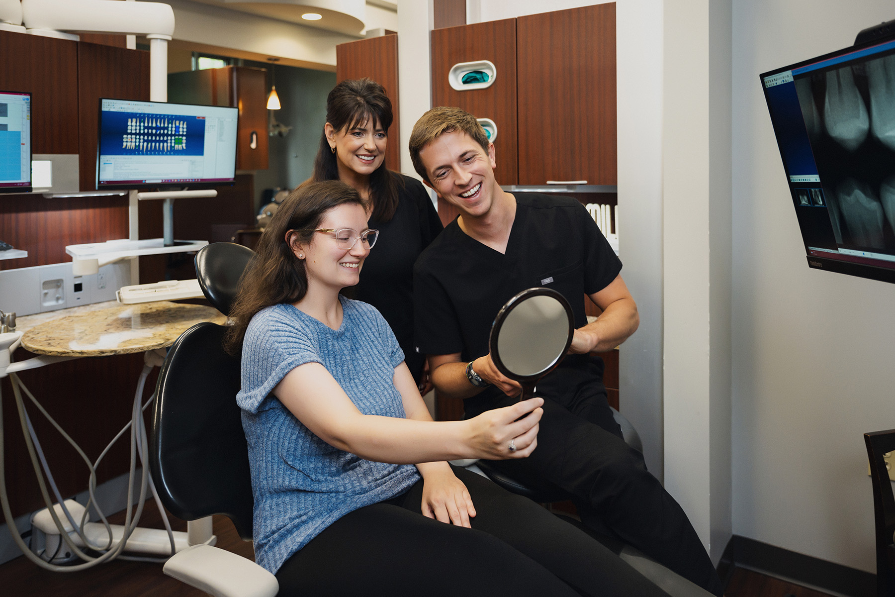Smiling dental patient holds mirror while two dental professionals look on in a modern clinic.