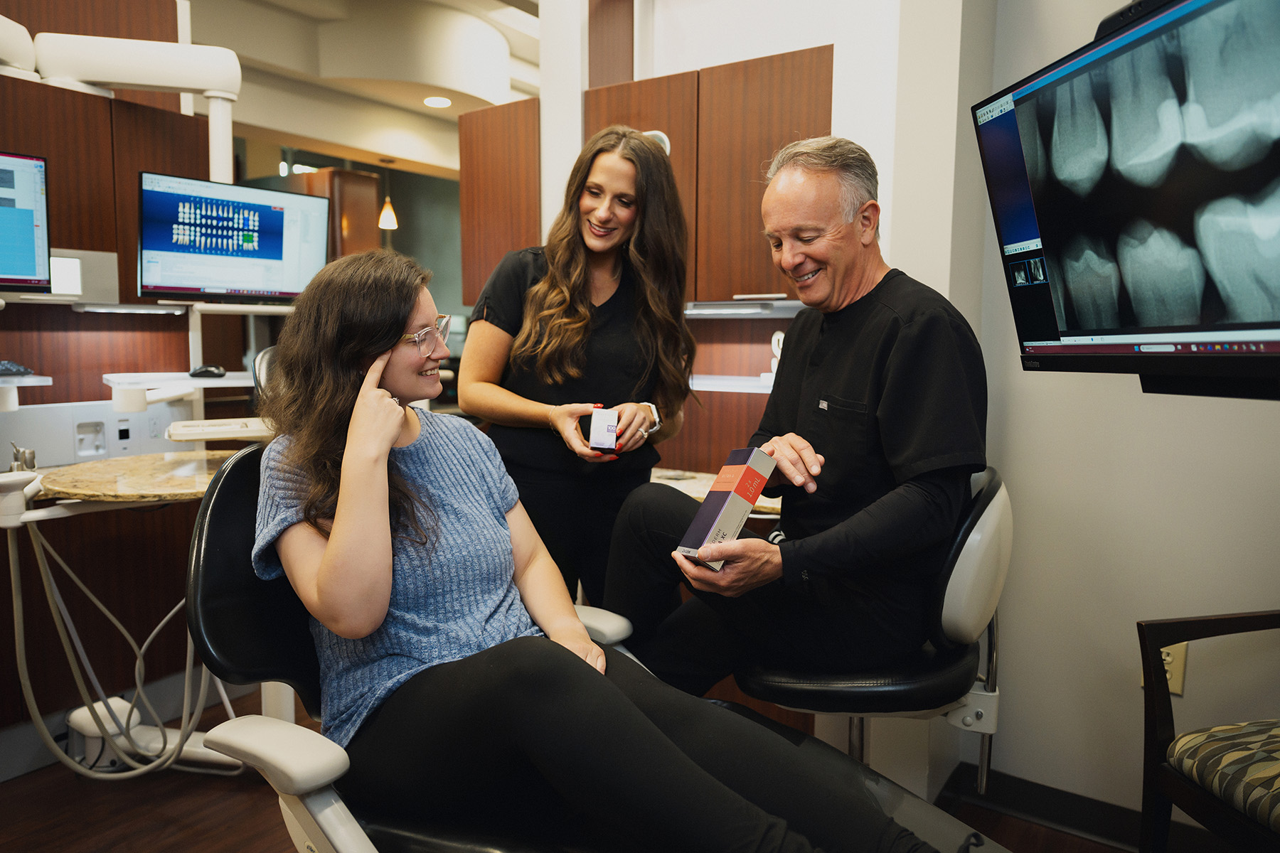 Dentist and assistant show dental product to female patient in a modern dental office with X-ray screen.