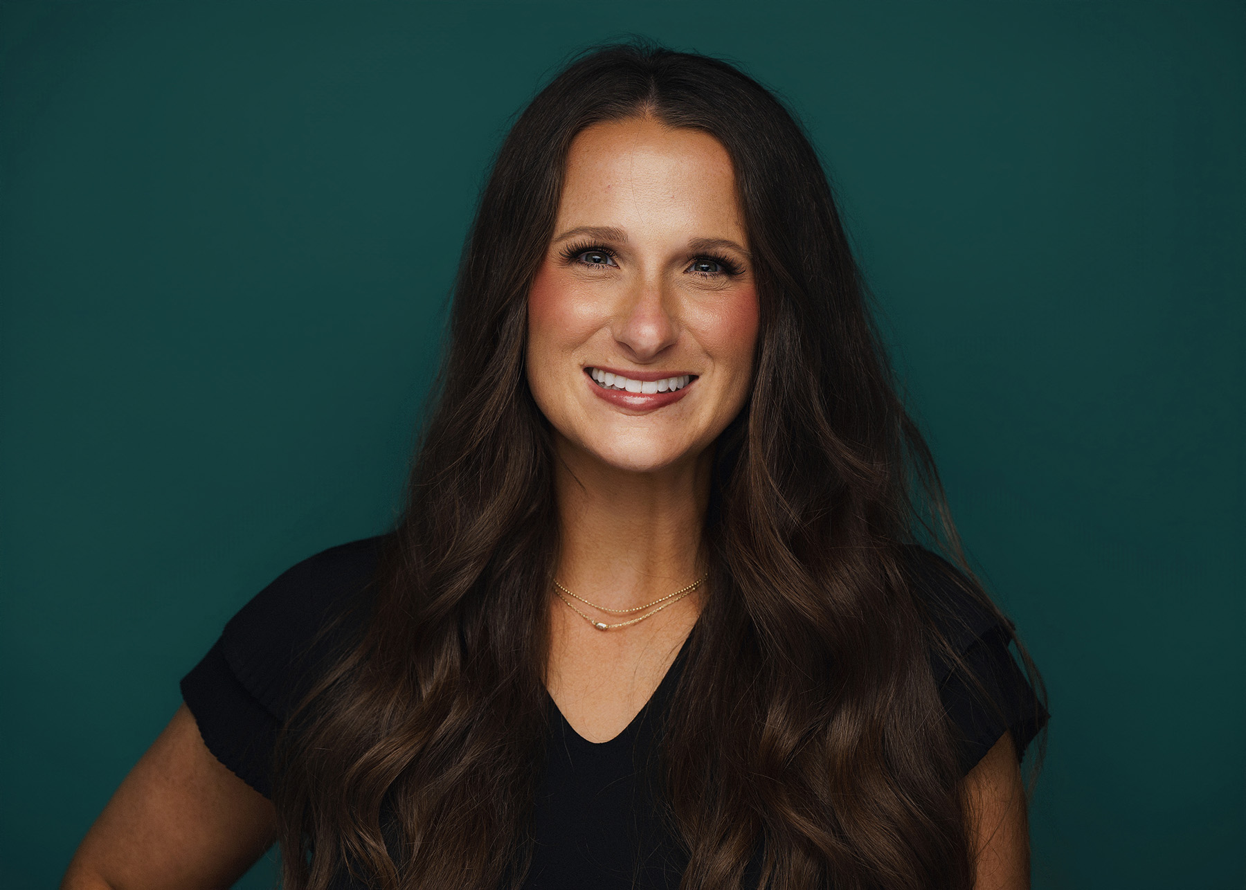 Smiling woman with long dark wavy hair wearing a black top and gold necklace on teal background.