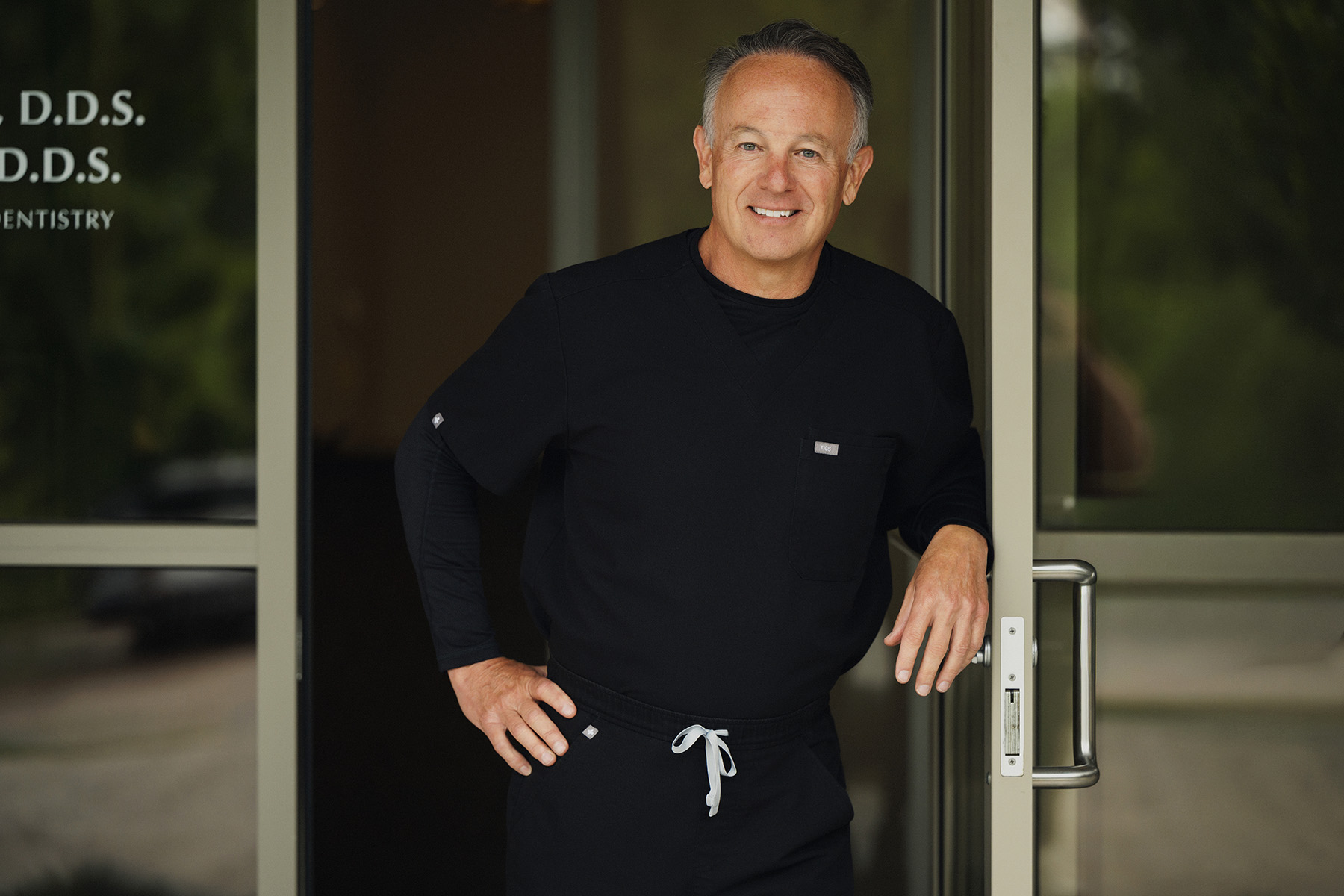 Smiling middle-aged man in black scrubs standing at a glass door with his hand on the handle.
