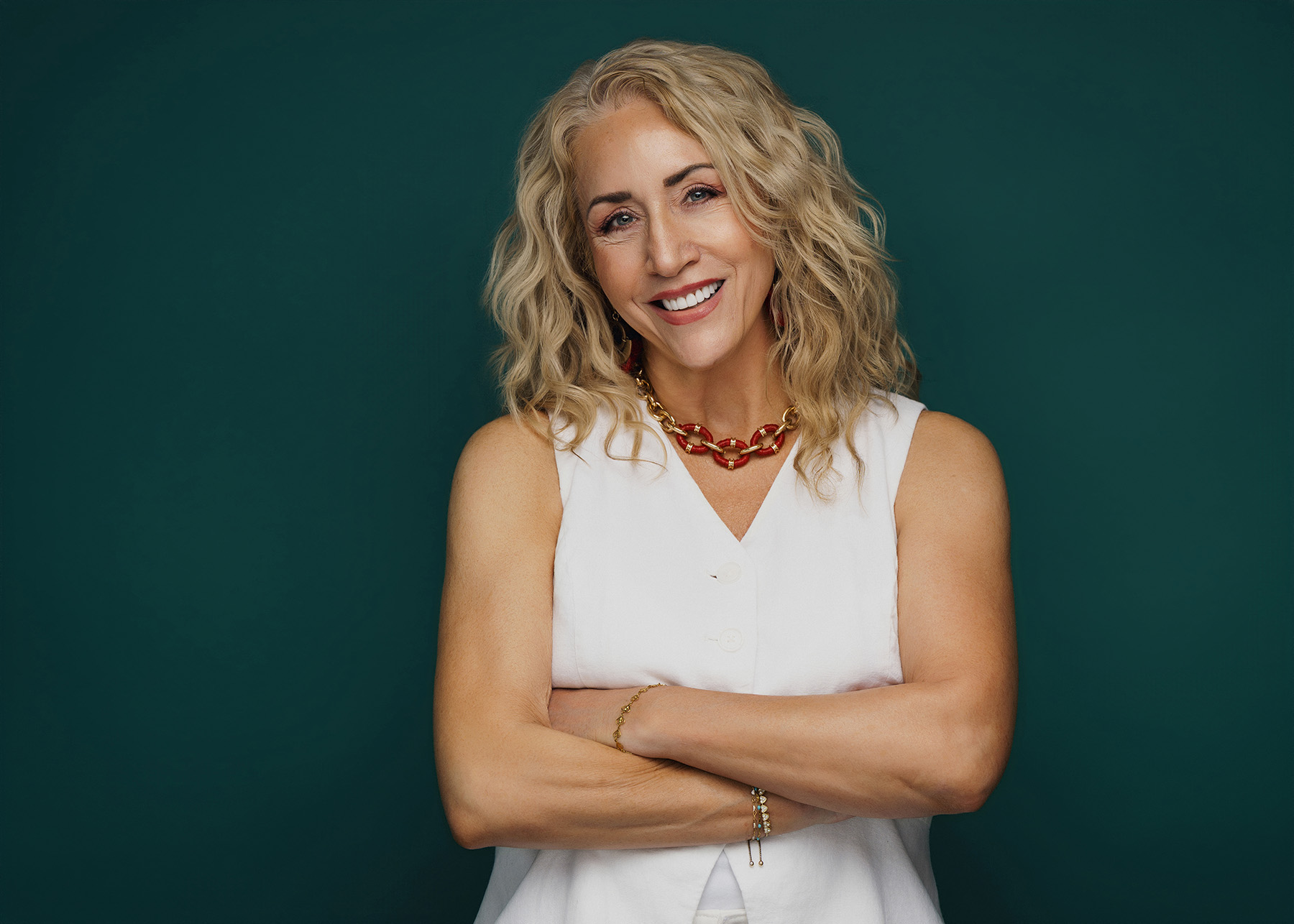 Smiling woman with curly blonde hair wearing white sleeveless top and red gold necklace over green background.