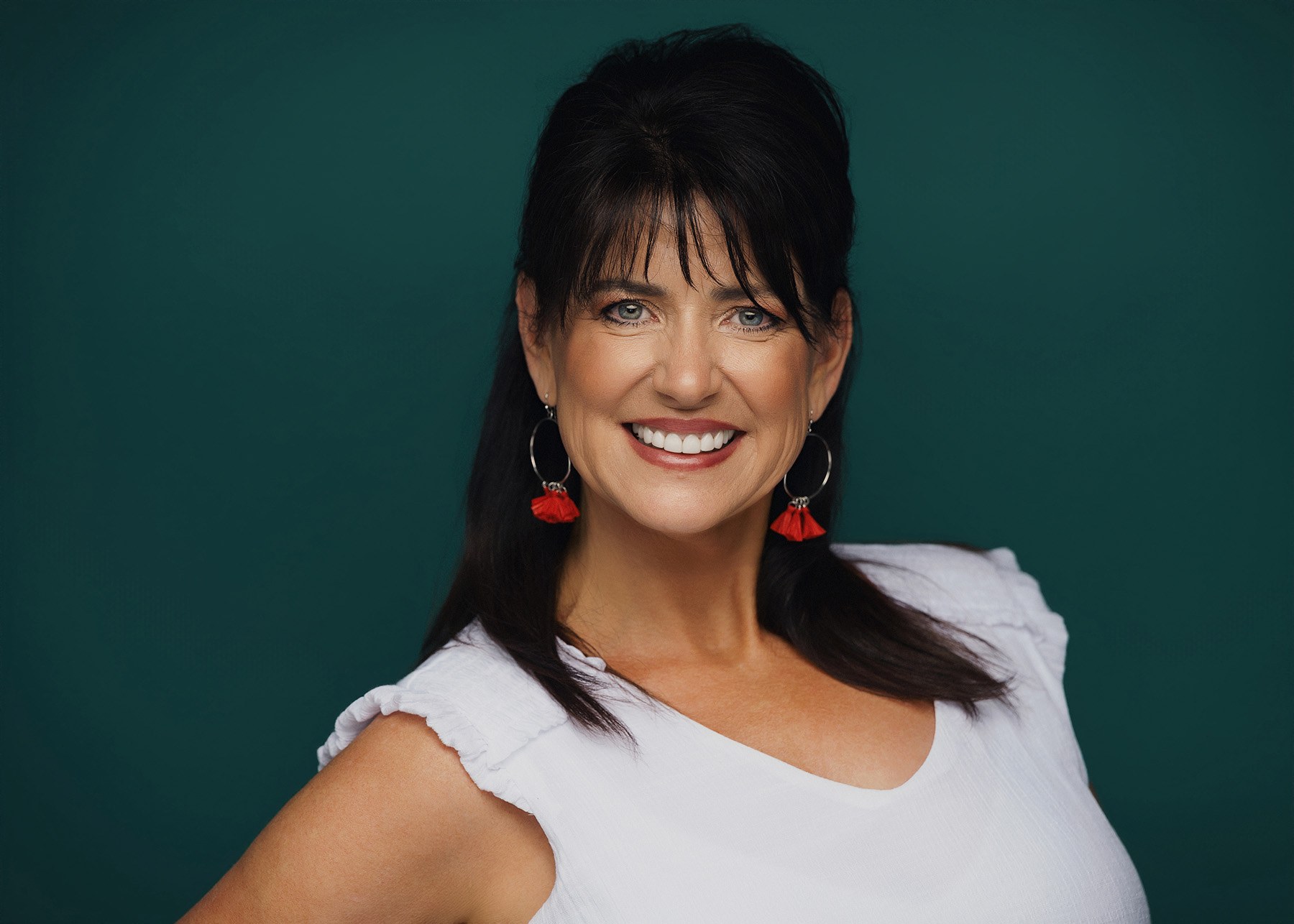 Smiling woman with dark hair wearing white top and red tassel earrings against teal background.
