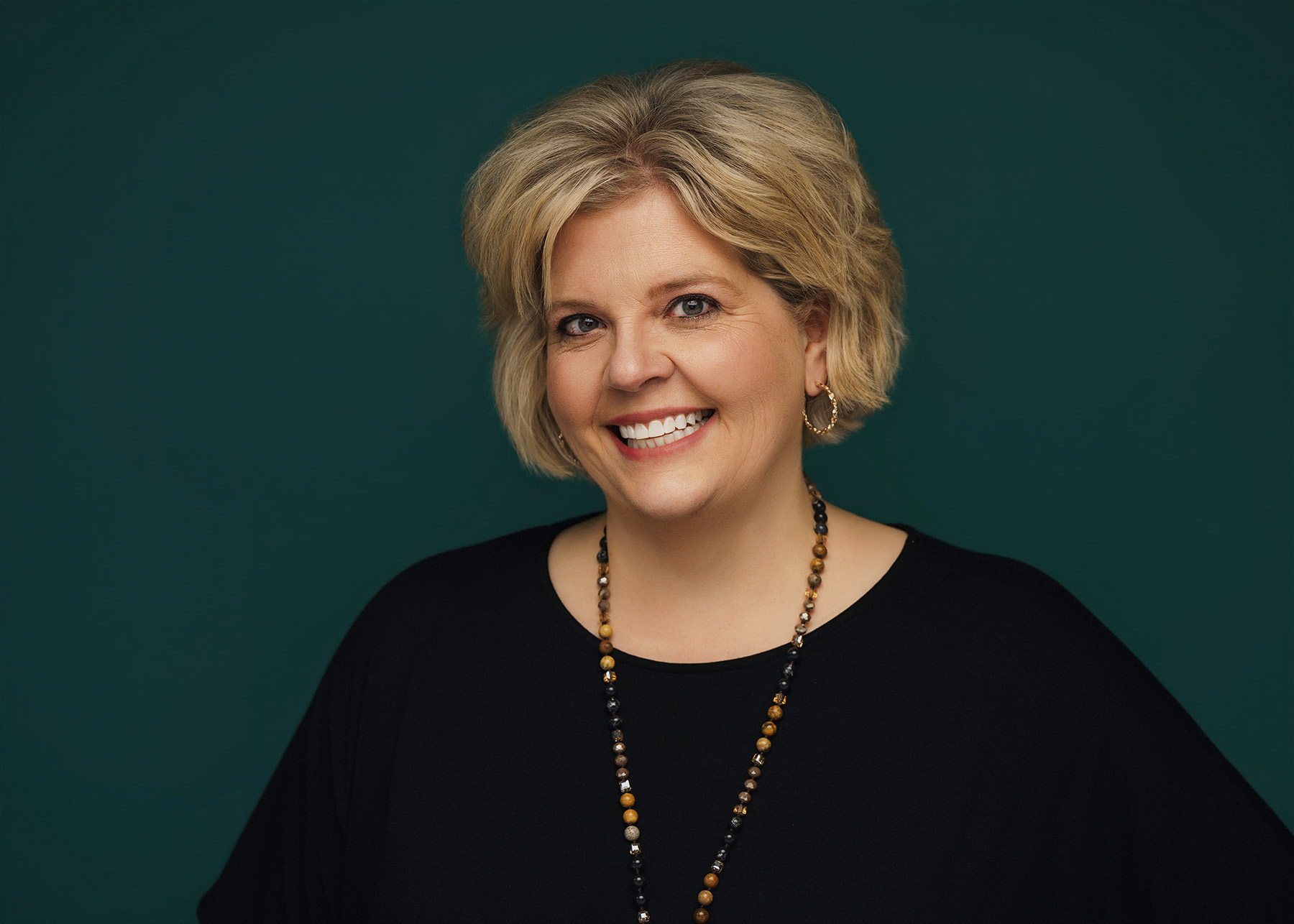Smiling woman with short blonde hair wearing hoop earrings and a beaded necklace against a dark background.