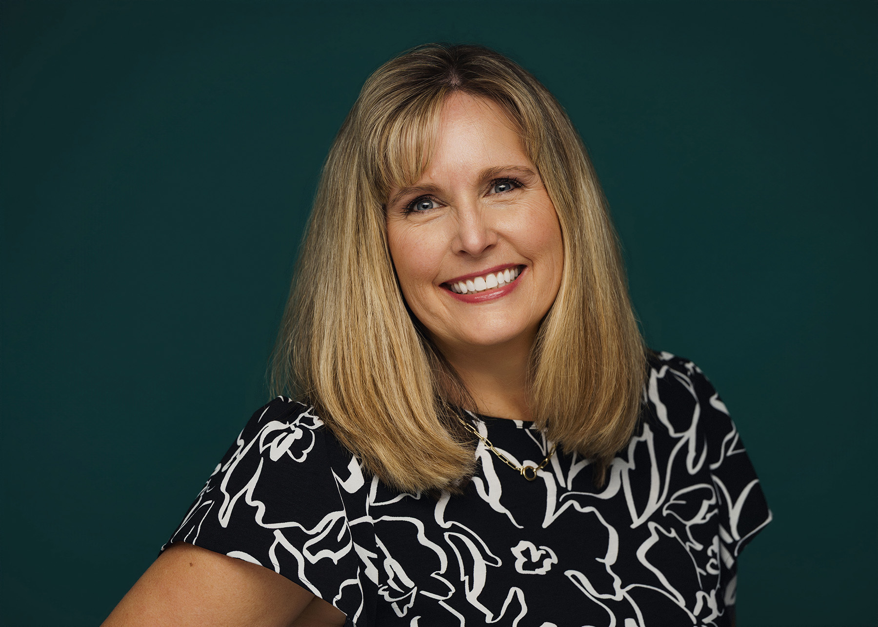 Smiling woman with shoulder-length blonde hair wearing a black and white floral top against a dark background.