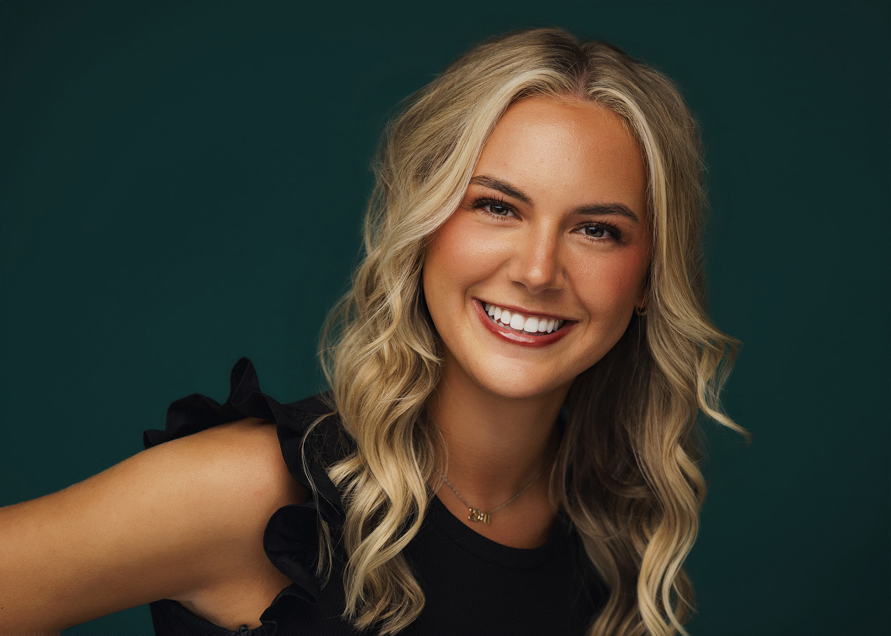 Smiling woman with blonde wavy hair wearing a black ruffled top and gold necklace against green.