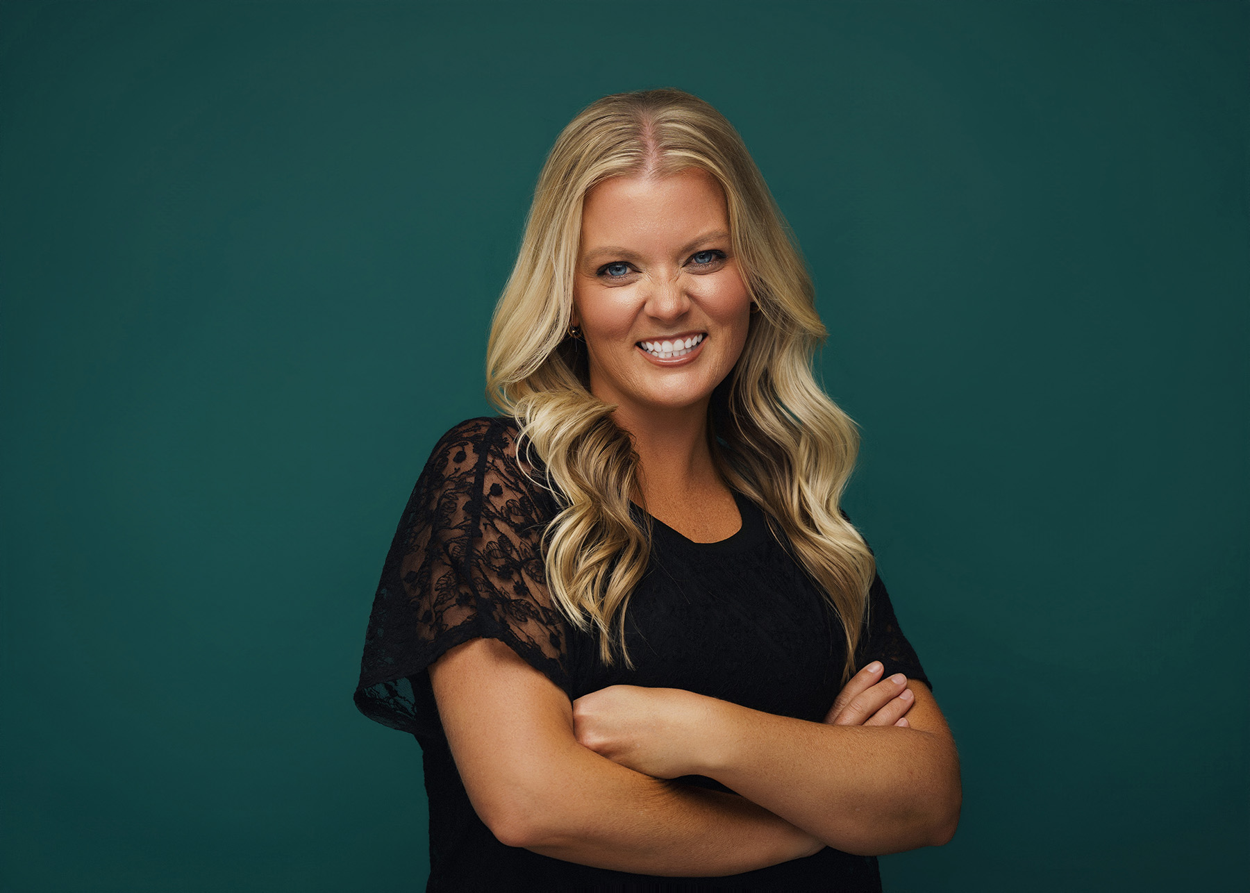 Blonde woman smiles with arms crossed against a dark green background wearing a black lace top.