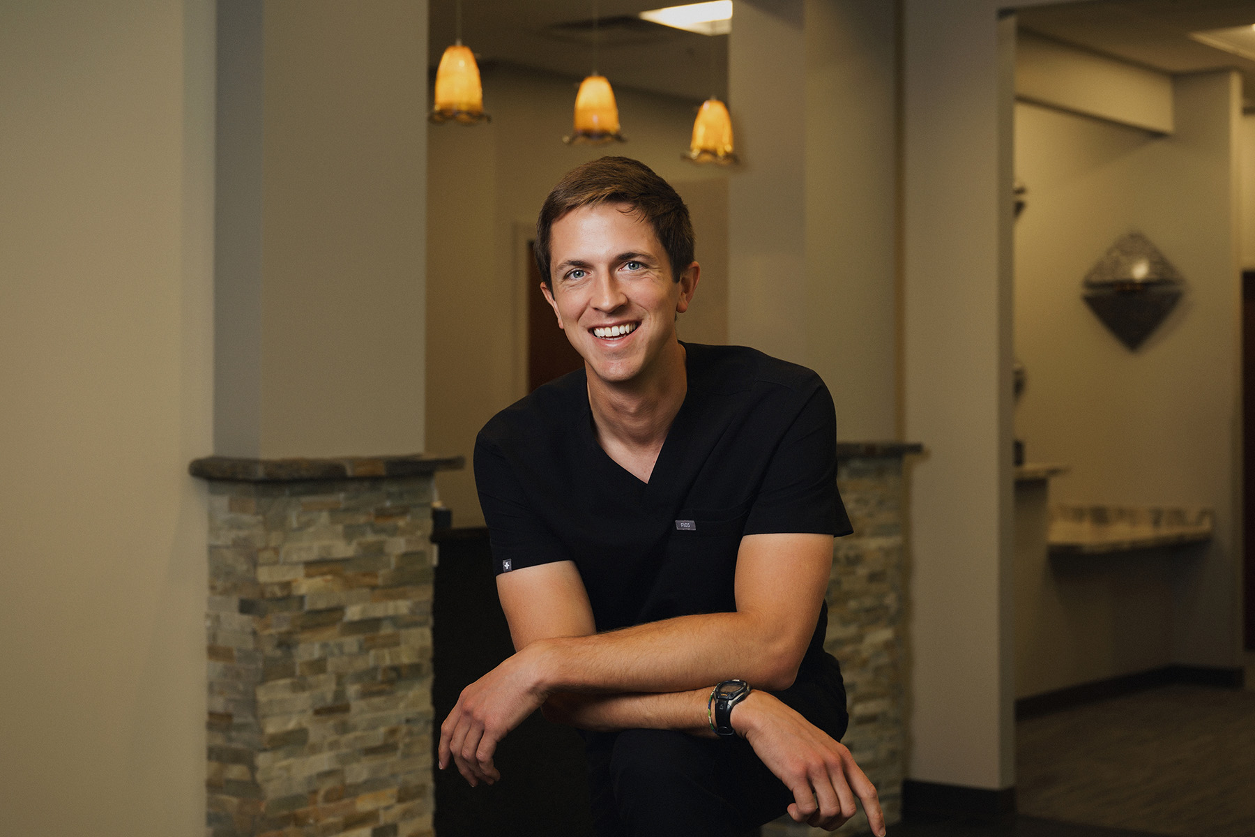 Smiling man in black scrubs seated indoors with stone wall and pendant lights behind him.
