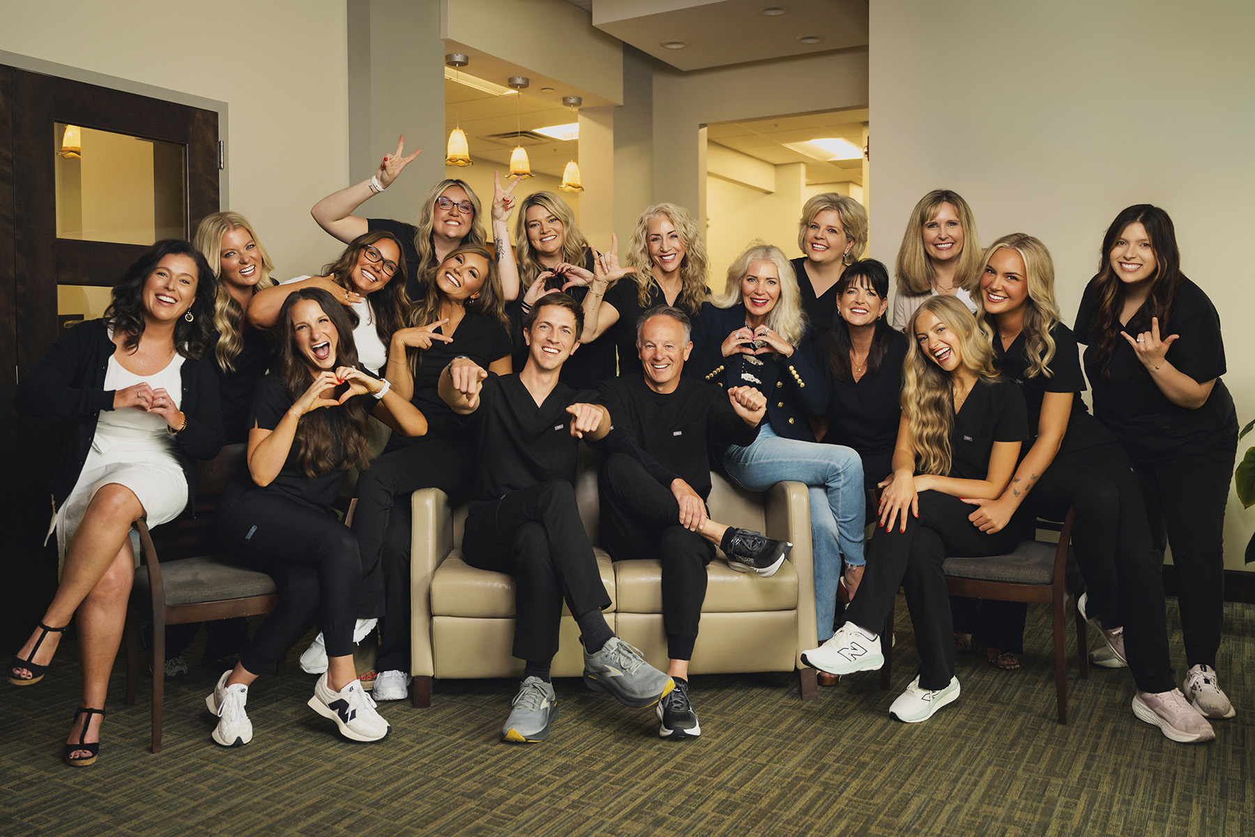 Group of smiling people, mostly women, posing indoors with hand heart and peace signs, two men seated front.