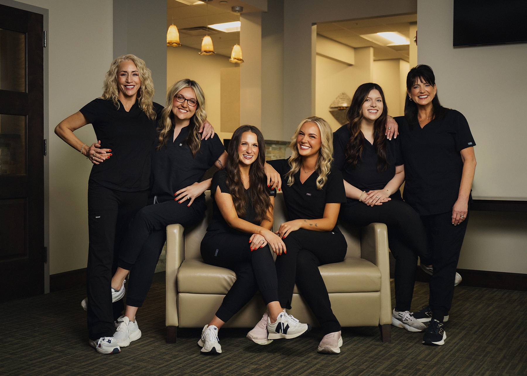 Six women in black scrubs smiling indoors, three seated on a beige sofa and three standing behind it.
