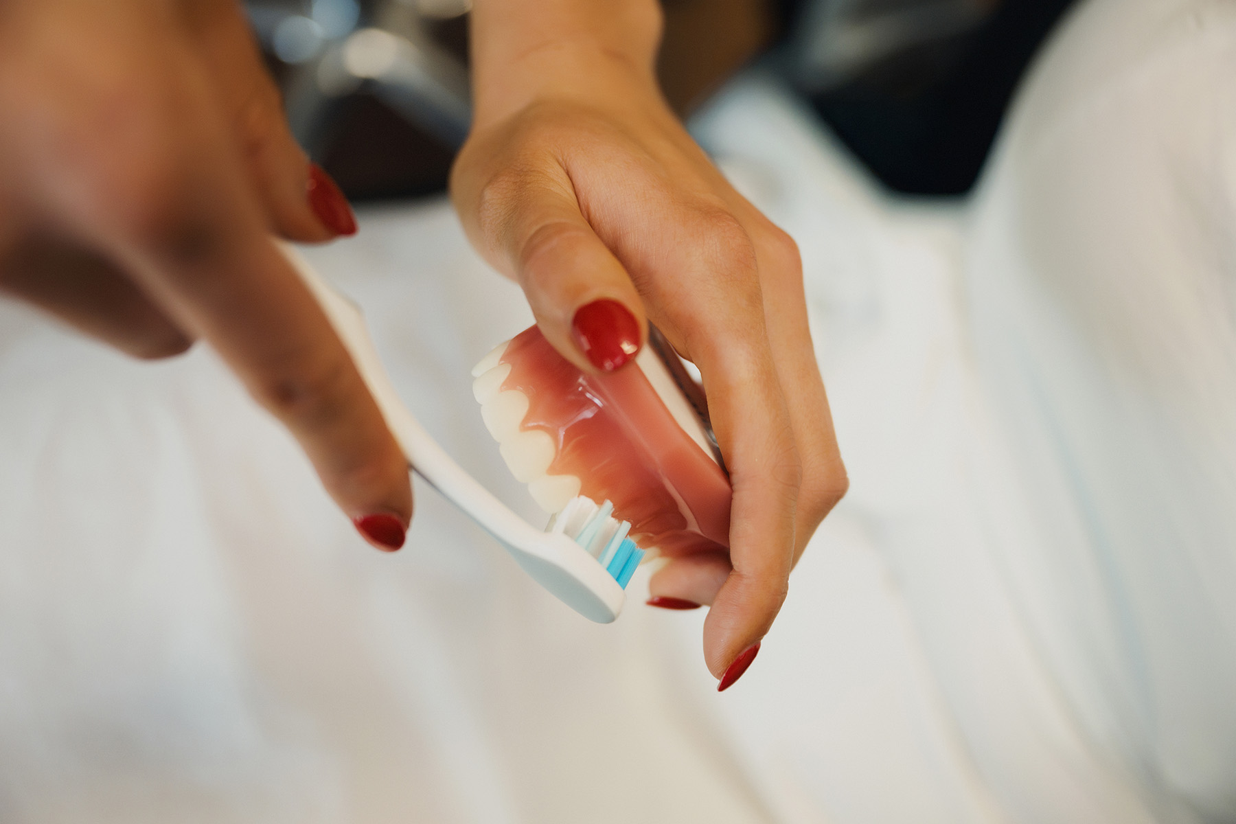 Hands with red nail polish holding dentures and brushing the teeth with a toothbrush.