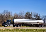 Blue semi-truck with a long silver tanker trailer parked on grass against a clear blue sky.