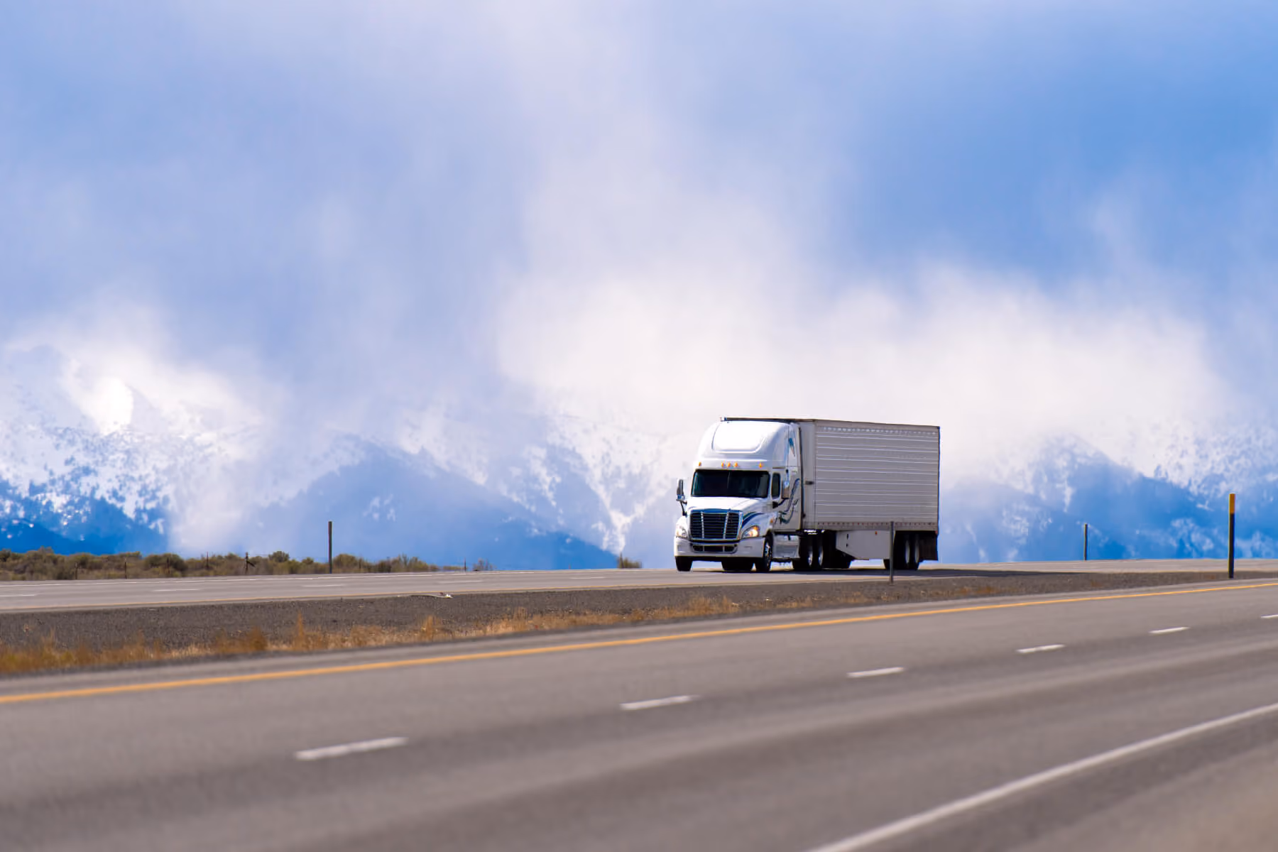 White semi-truck driving on highway with snow-covered mountains and cloudy sky in the background.