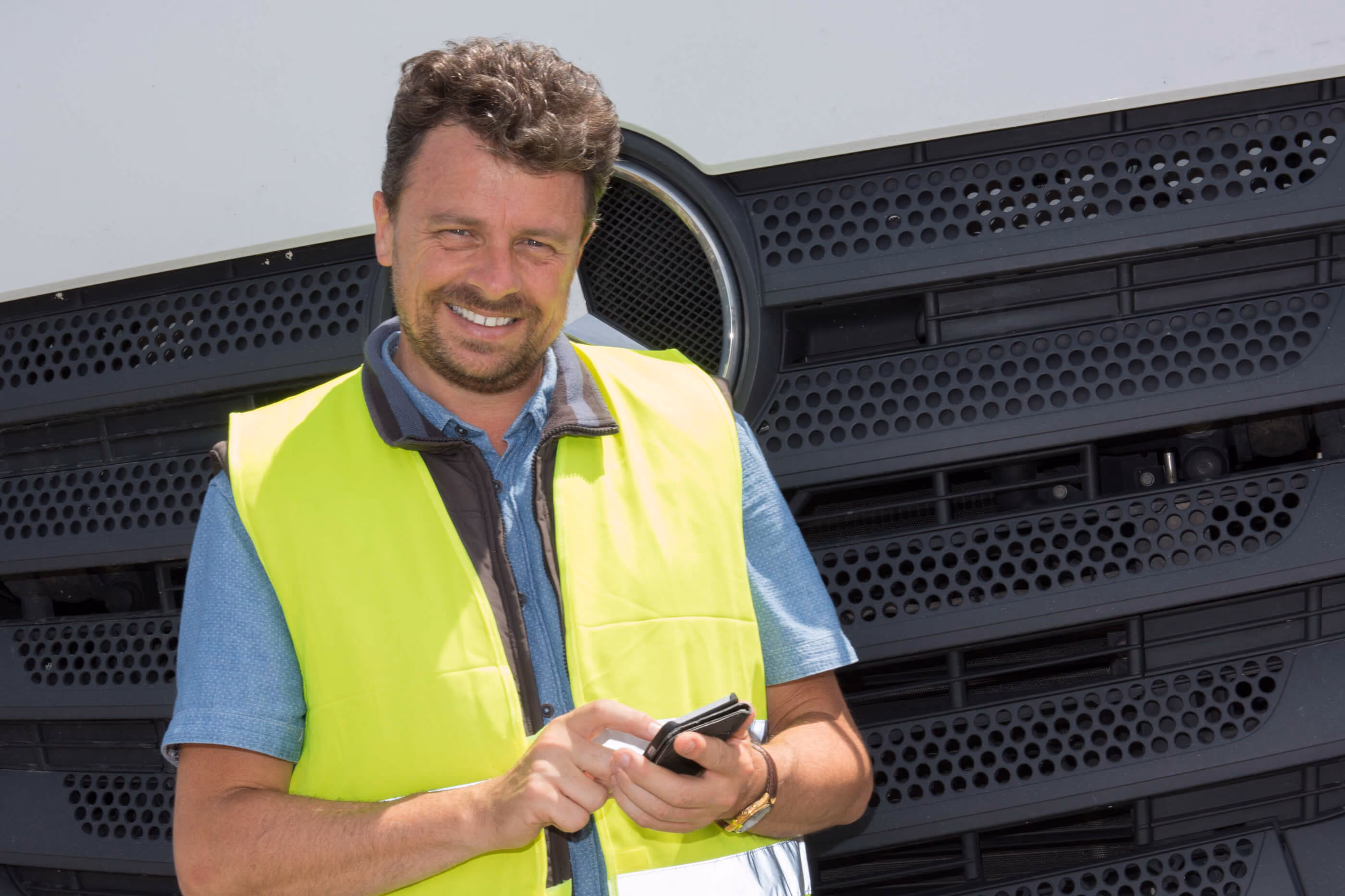 Smiling man in yellow safety vest using smartphone, standing in front of a large truck grille.