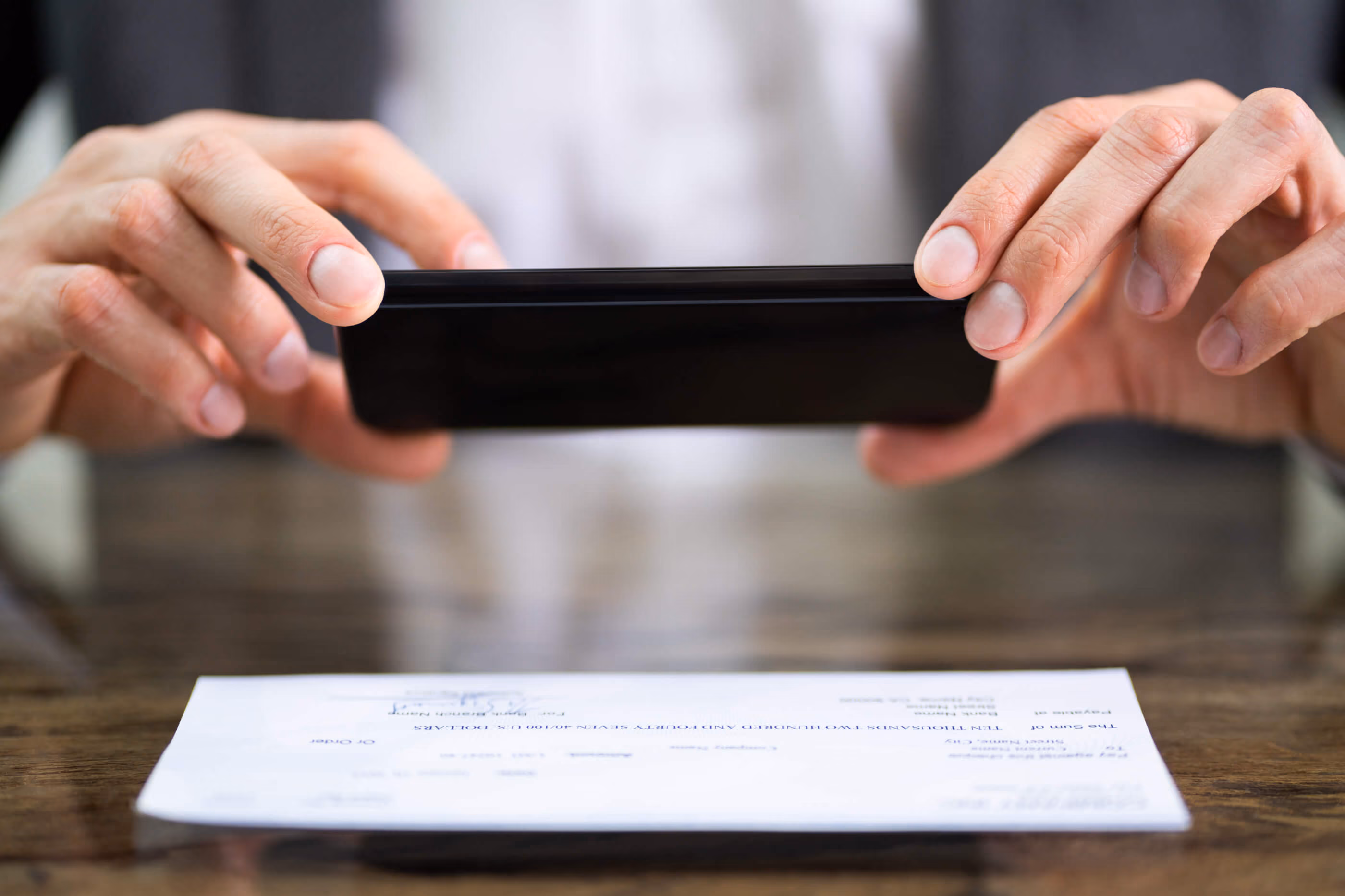 Close-up of hands holding a smartphone to take a photo of a check on a wooden table.