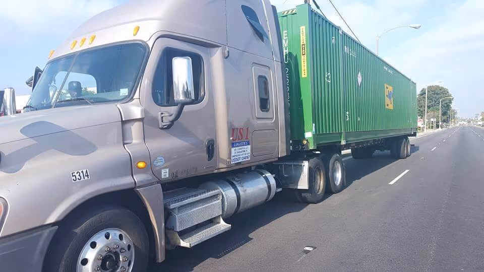 Pewter-colored semi-truck with a green shipping container driving on a wide road in daylight.