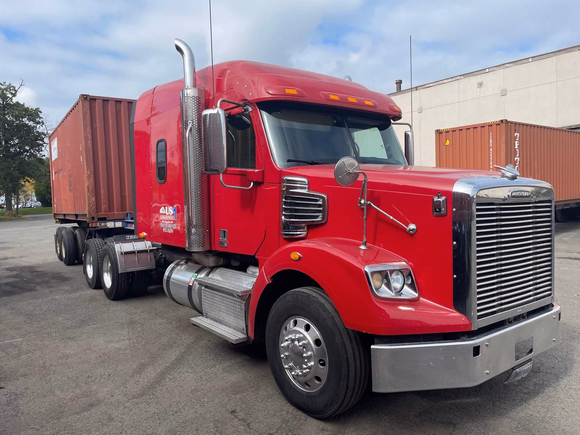 Red Freightliner semi-truck with attached shipping container parked on paved surface near warehouse.