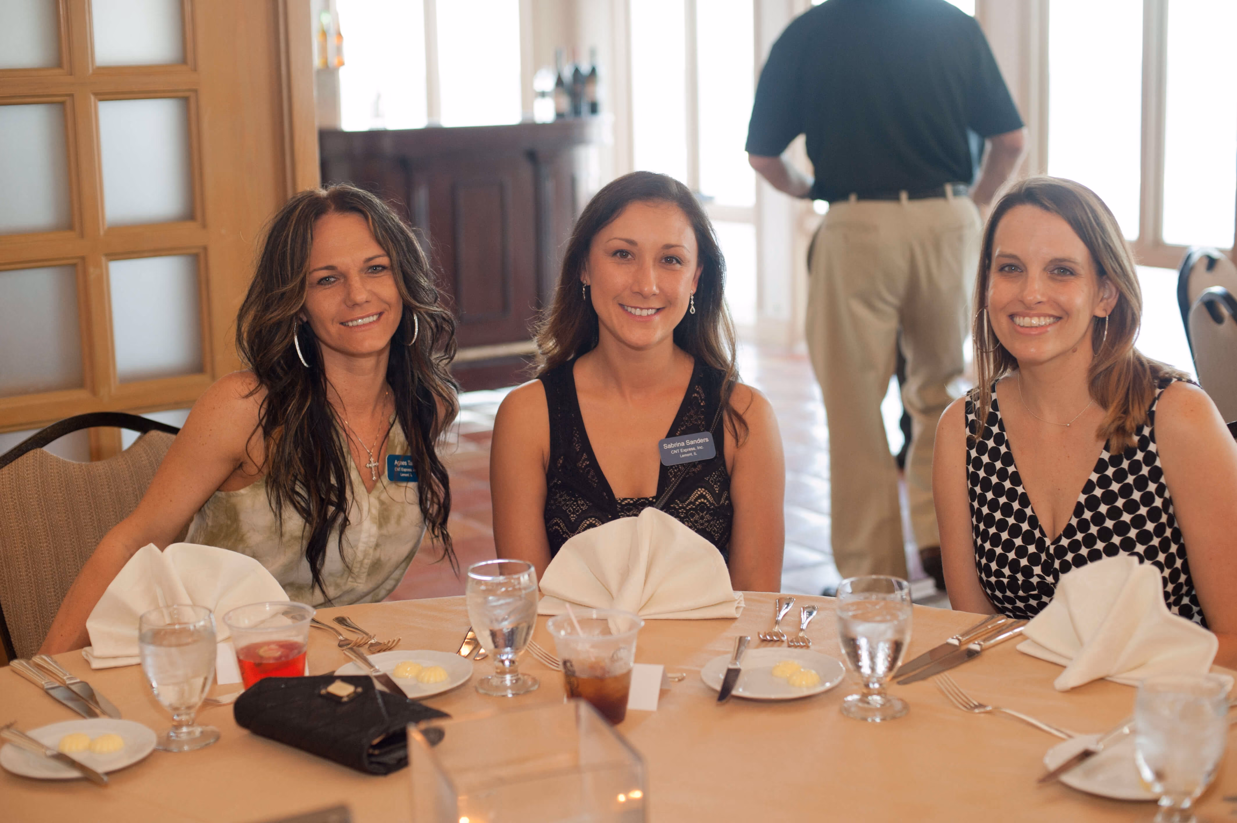 Three women seated at a round table set with glasses, plates, and napkins, smiling at the camera.