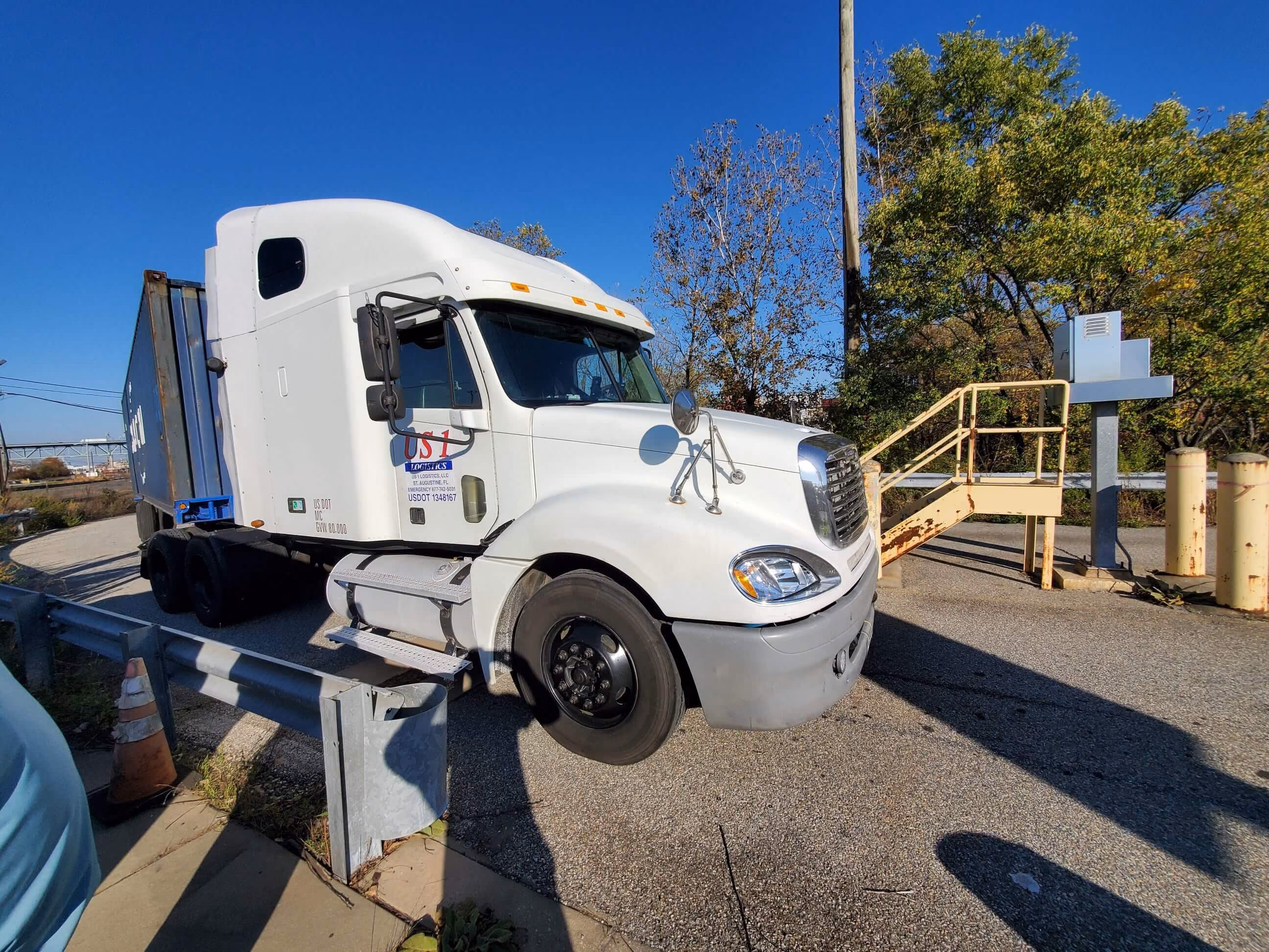 White semi-truck with a blue trailer parked near a rusty yellow metal staircase and trees.