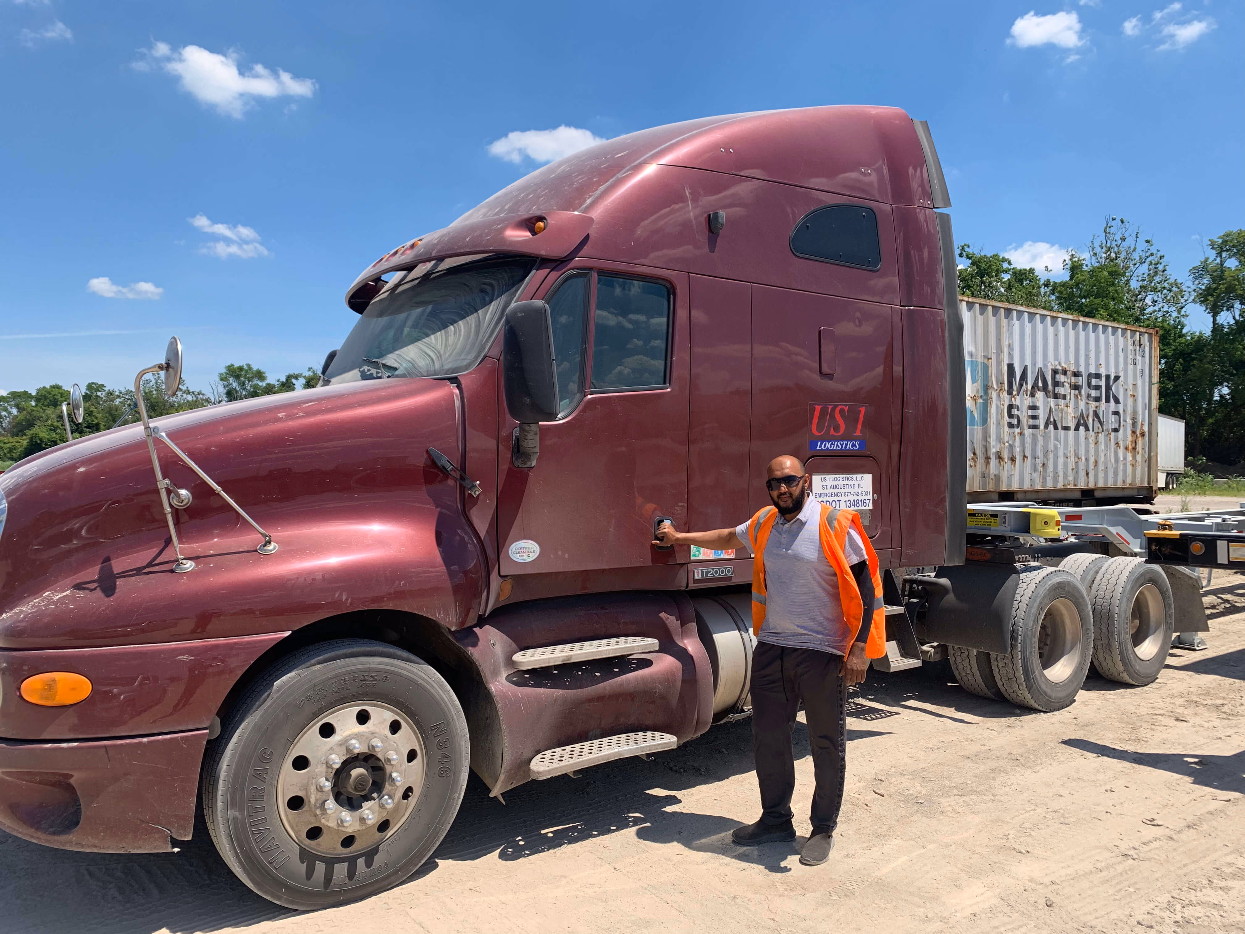 Man wearing an orange safety vest standing by the door of a maroon semi-truck with a shipping container trailer.