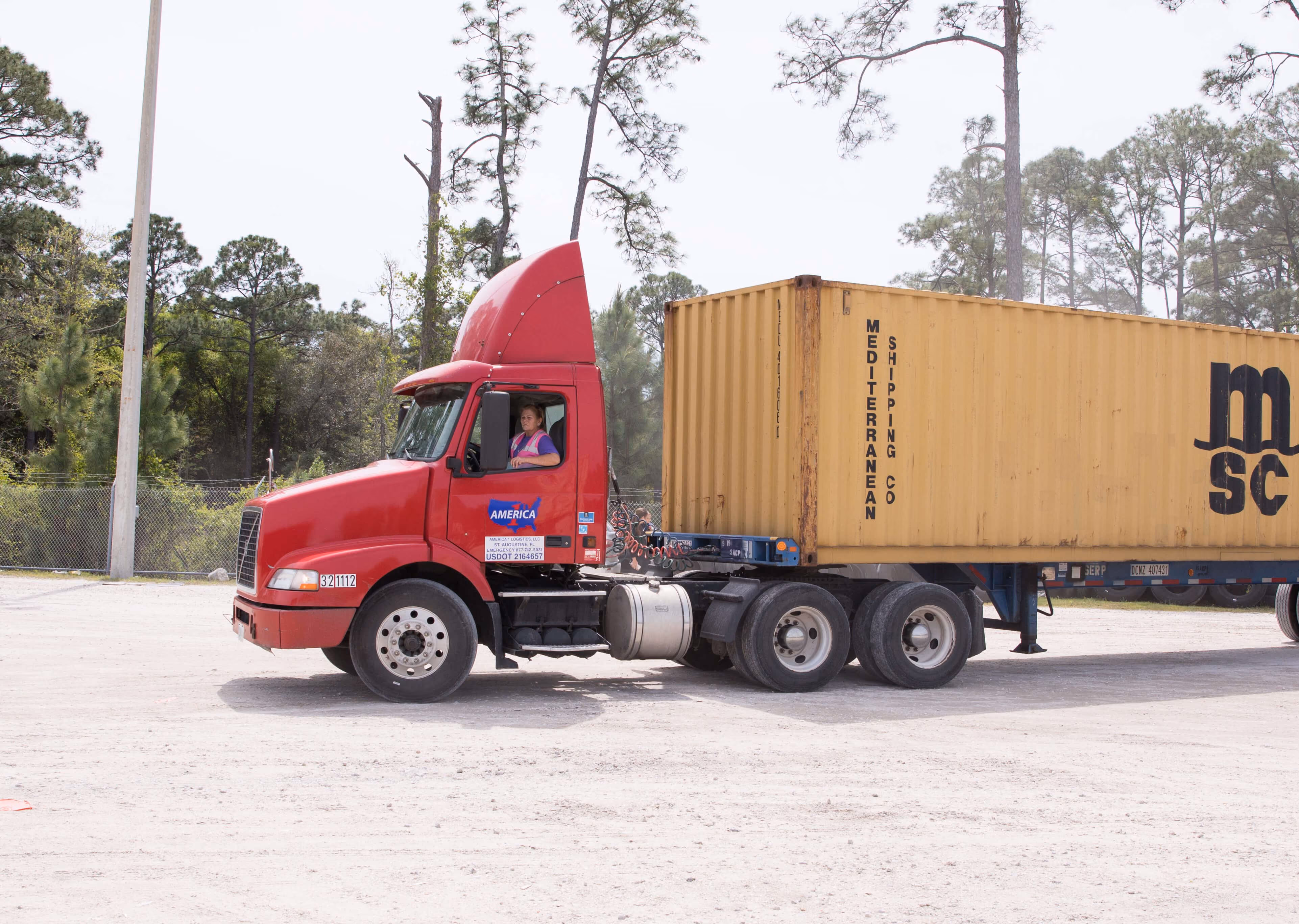 A woman in a pink safety vest sits in the cab of a red semi truck attached to a yellow shipping container.