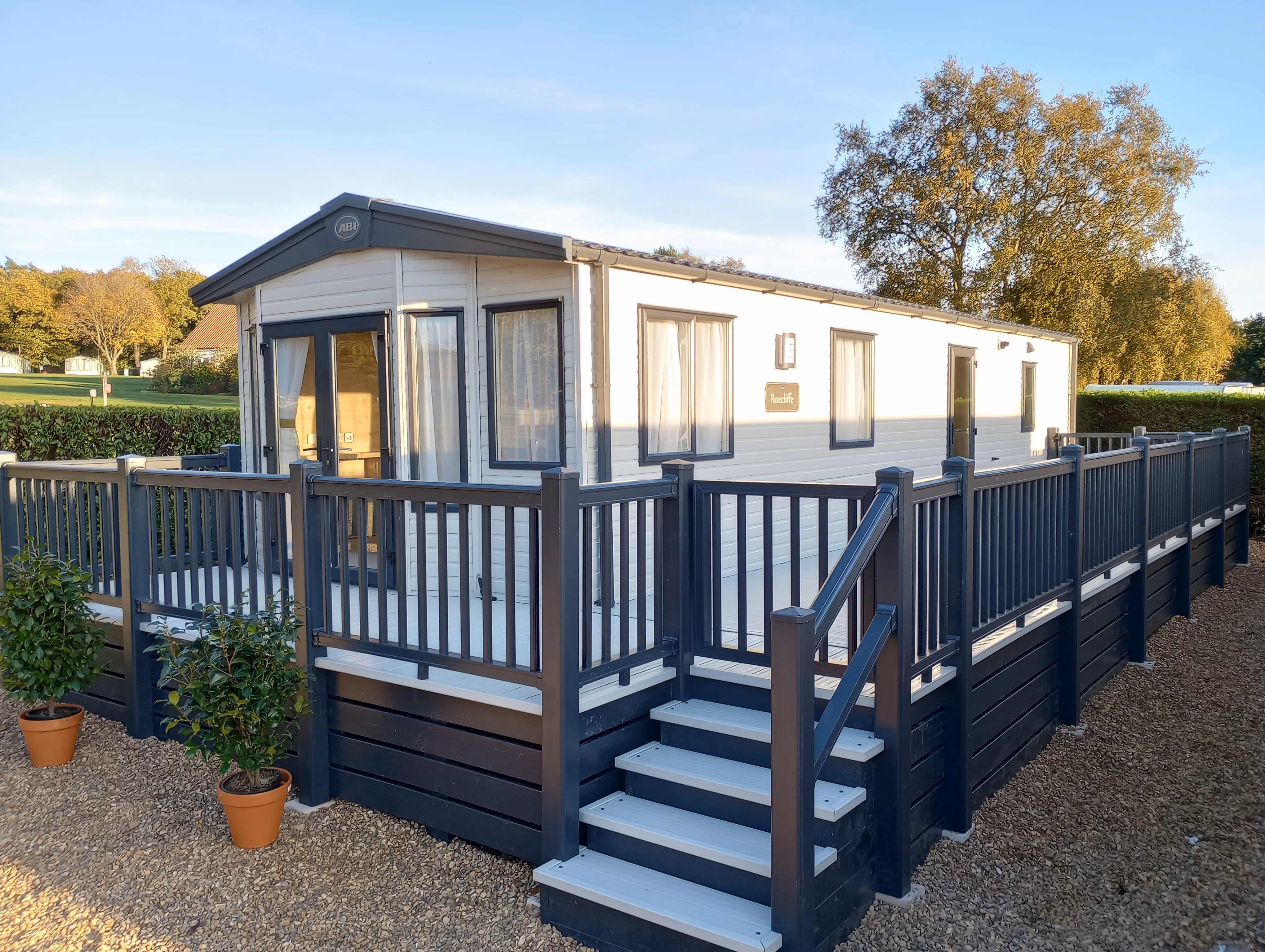 White mobile home with dark blue railing and steps on a gravel path with potted plants nearby.