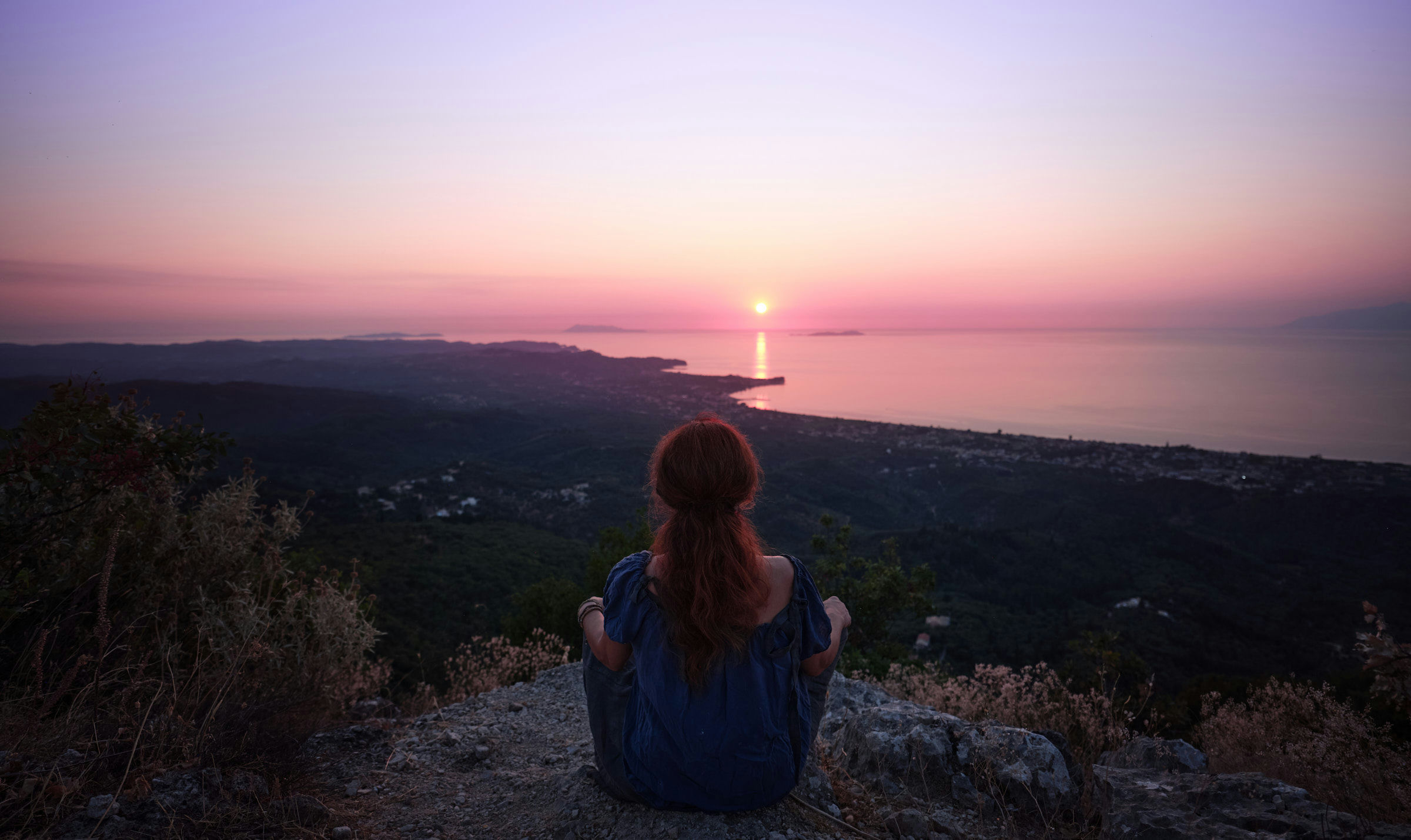 Erin rothaarige Frau sitzt auf einem Felsen und blickt in den Sonnenuntergang über dem Ionischen Meer
