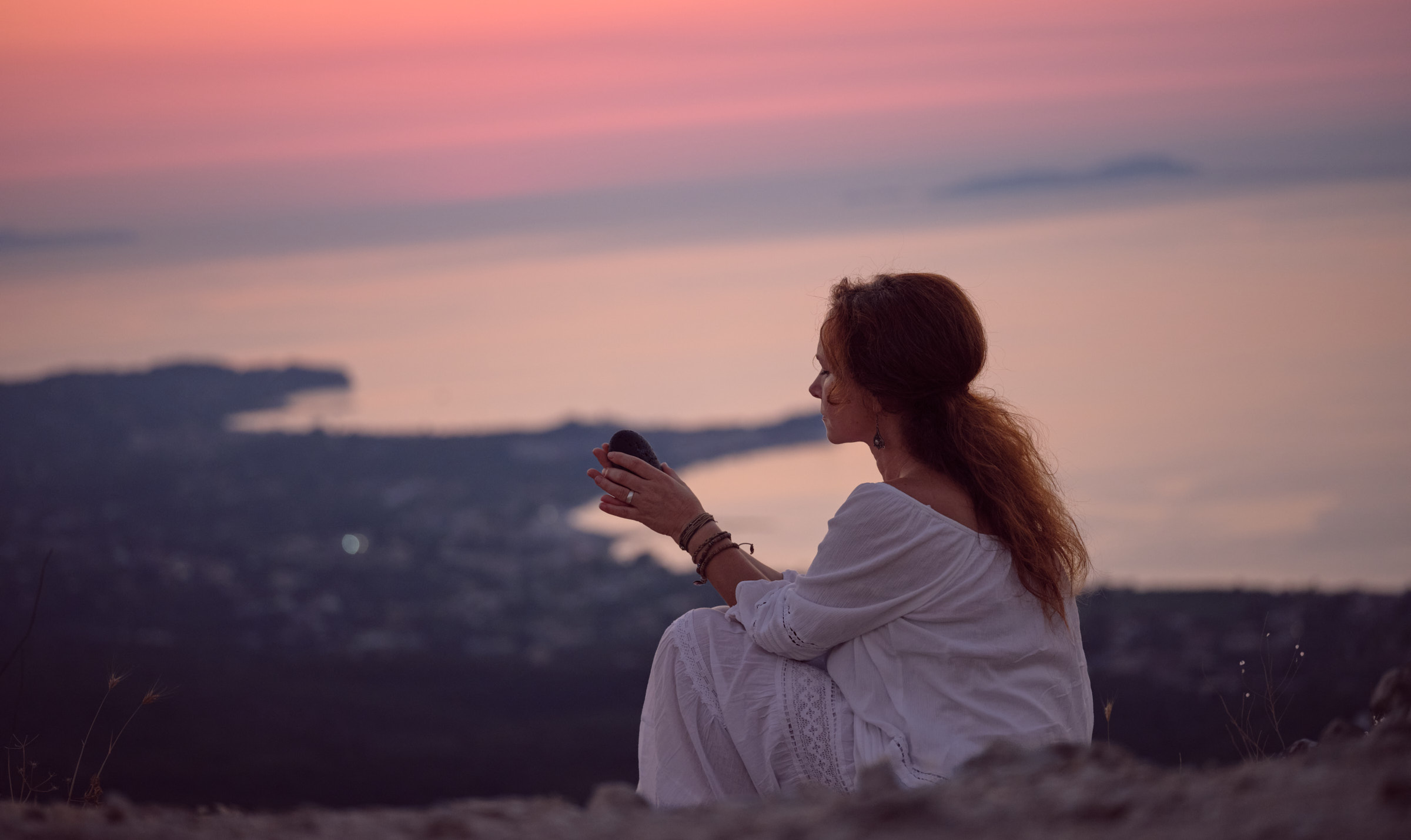 Eine Frau sitzt im lila farbigen Zwielicht auf einem Berg über dem Meer - in ihrer Hand hält sie einen schwarzen Stein