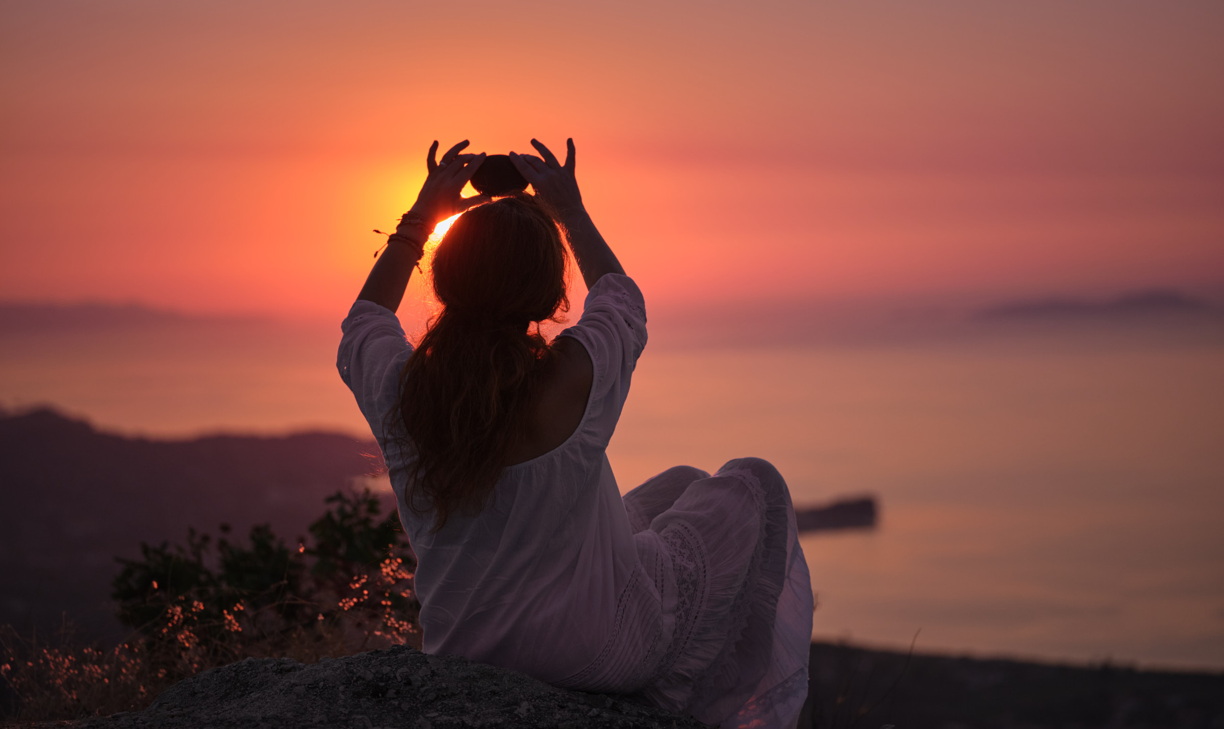 Eine Frau, das Meer überschauend, hält einen Stein in die untergehende Sonne