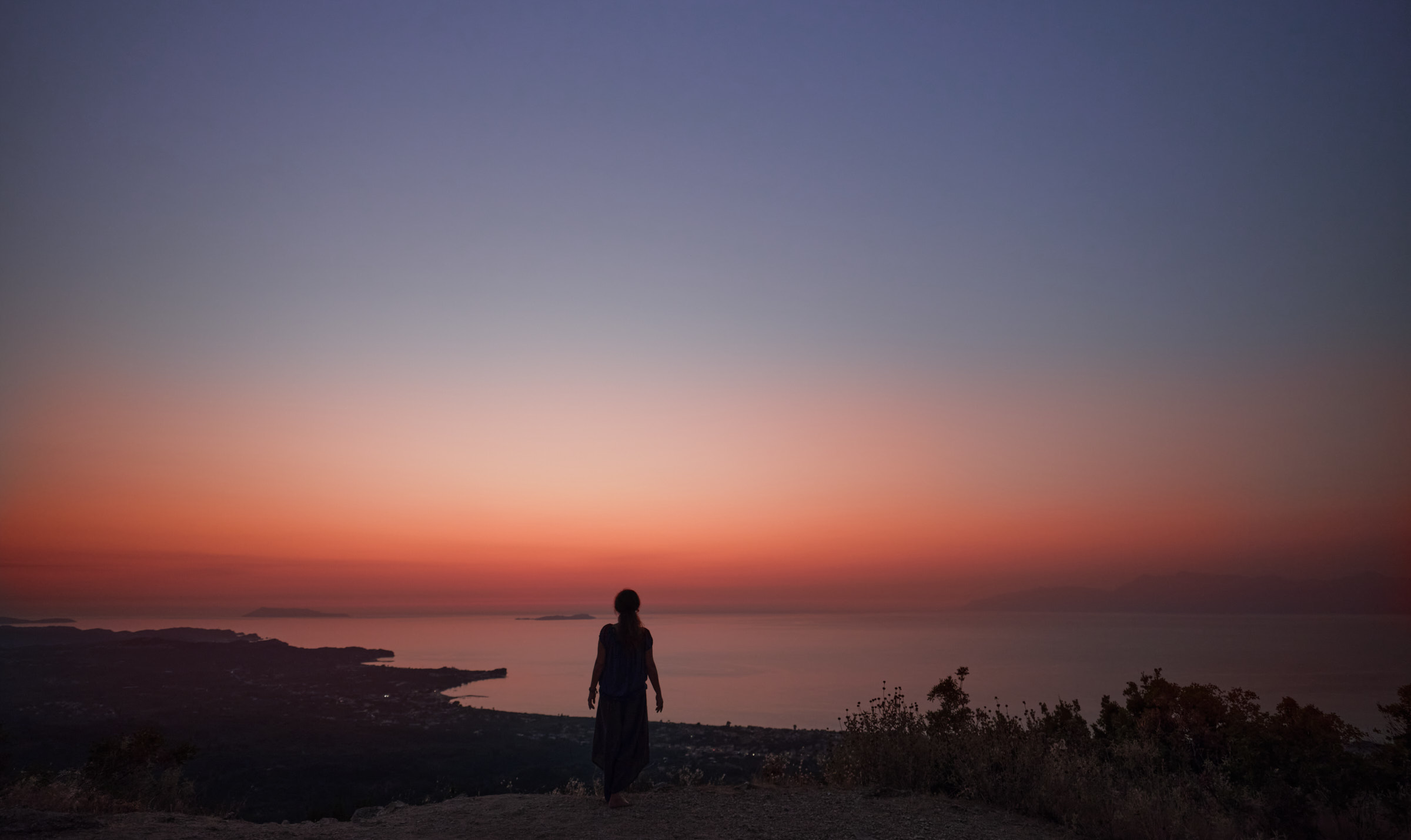 Eine Frau steht auf einem Berg, vor ihr das Panorama eines spätabendlichen Himmels über dem Ionischen Meer.