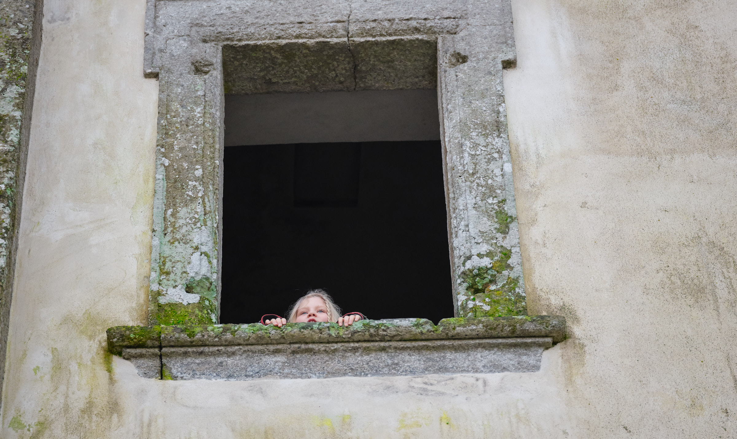 Ein kleines Mädchen blickt aus der steinernen Fensteröffnung eines alten, moosbewachsenen Hauses.