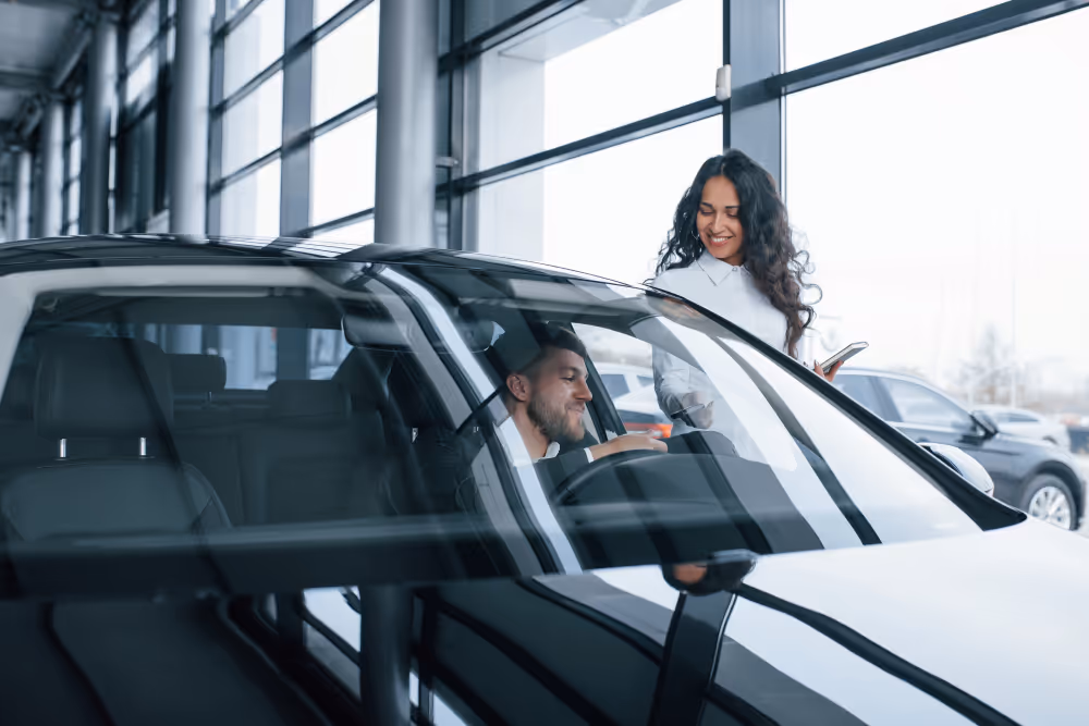 A woman standing outside is handing car keys to a man sitting inside a black car in a showroom.