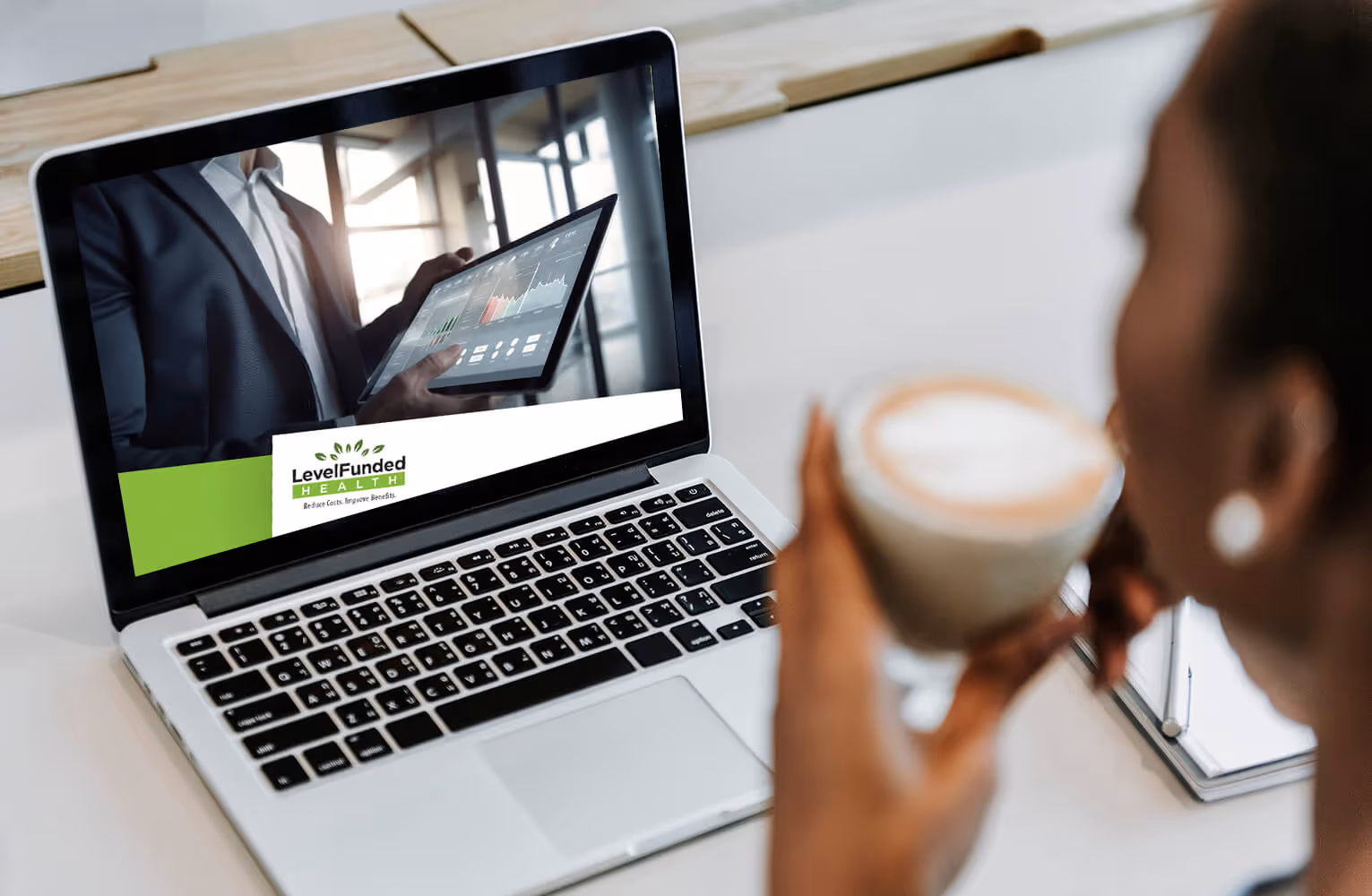 Person holding a coffee cup watching a webinar on a laptop showing a business presentation with charts and the LevelFunded Health logo.
