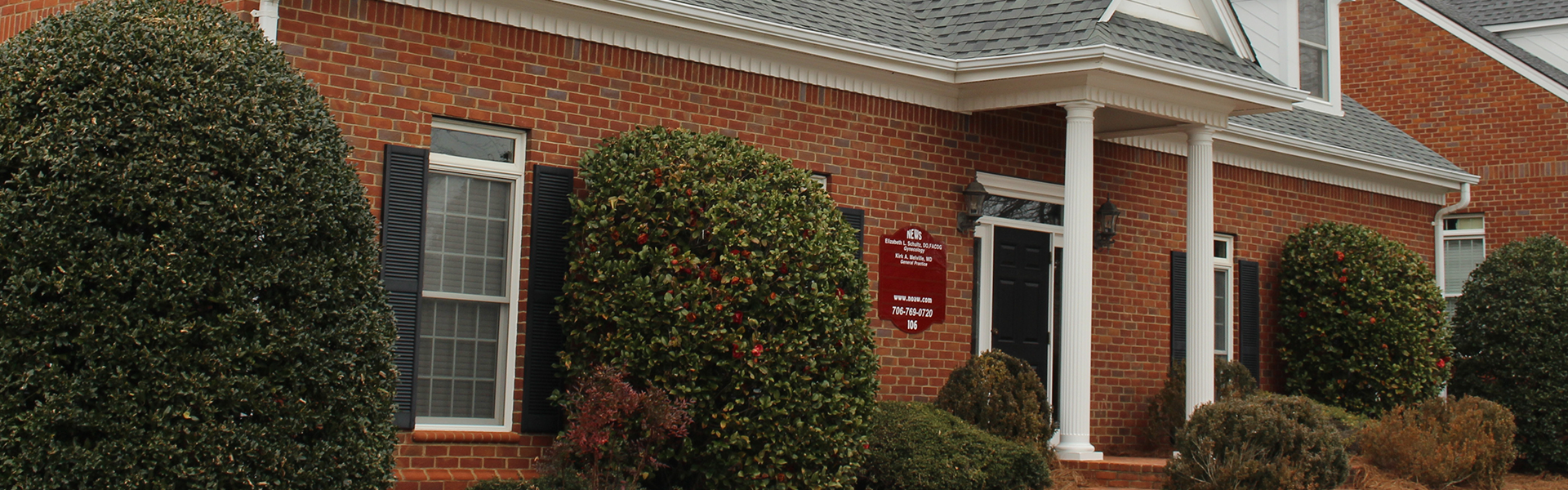 Brick building with black shutters, white columns, a black door, and neatly trimmed bushes and shrubs in front.