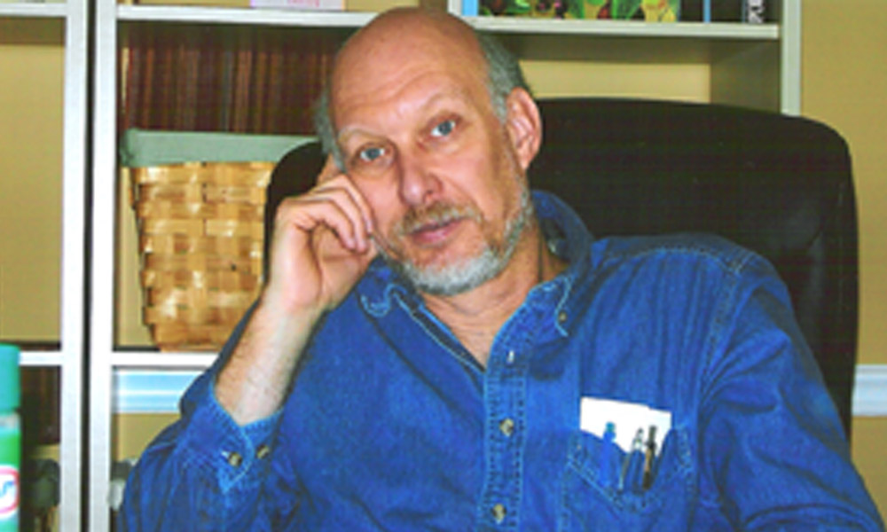 Middle-aged man with a beard wearing a blue shirt, sitting thoughtfully with his head resting on his hand in an office setting.