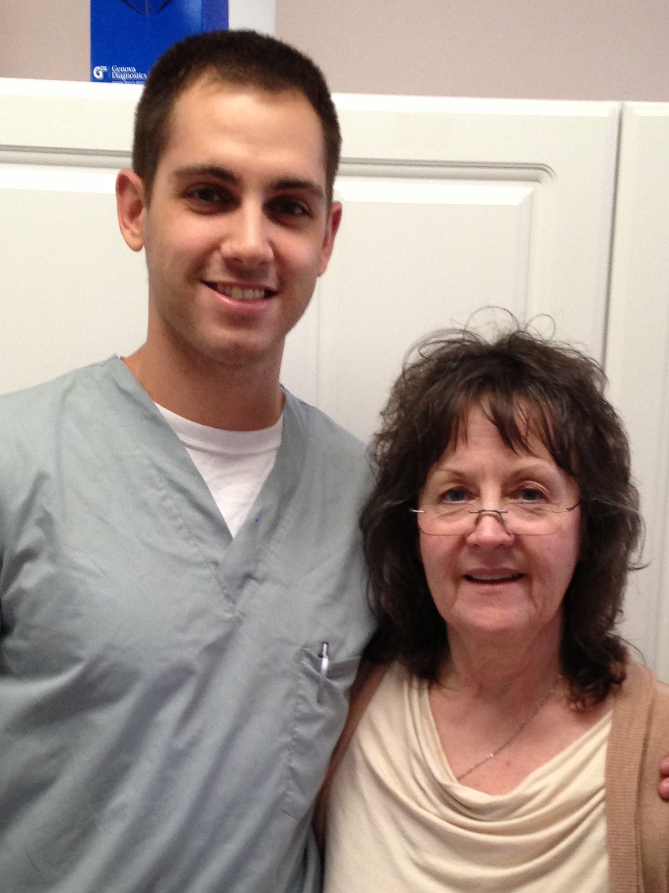 Young man in gray medical scrubs standing next to an older woman with glasses, both smiling.