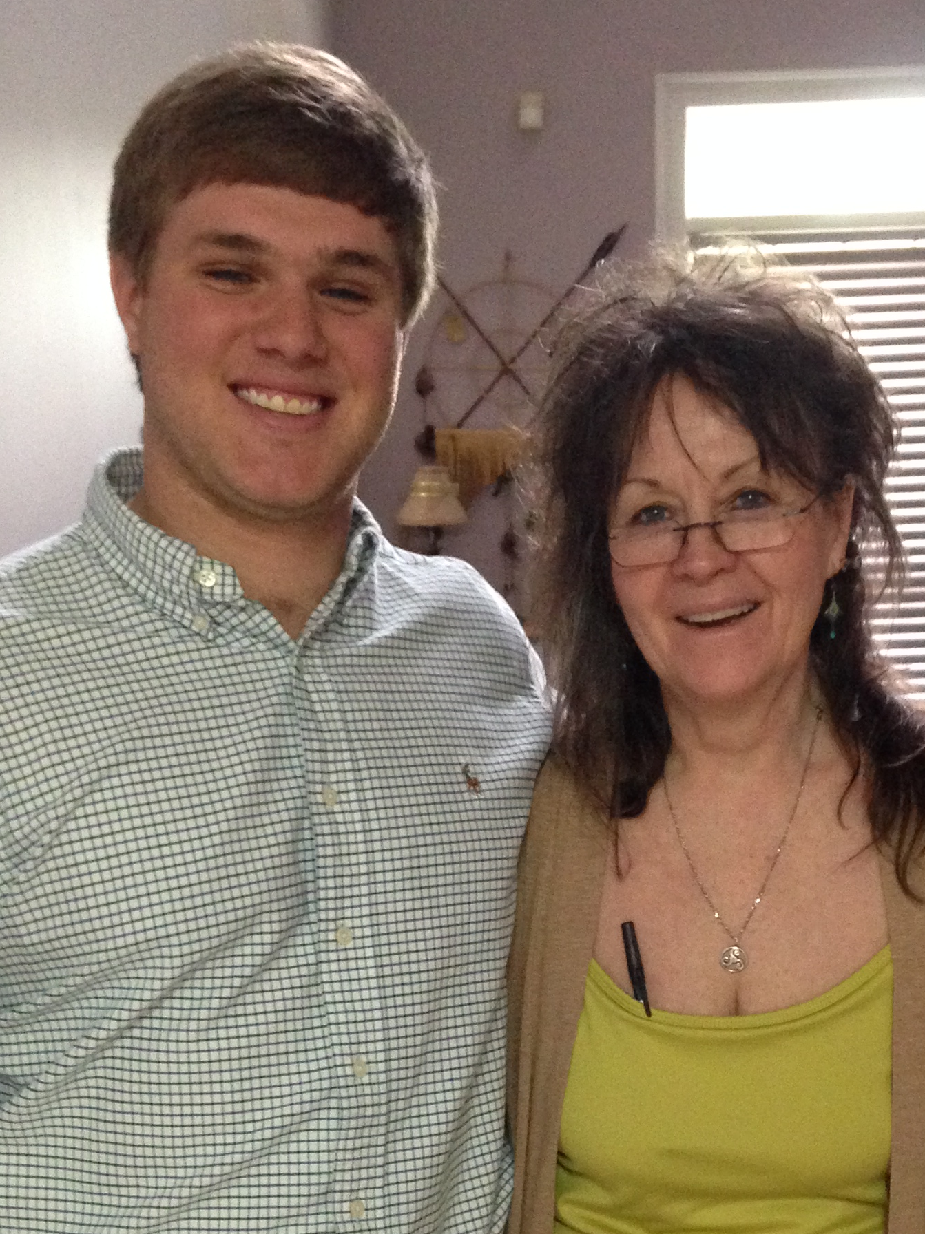 Smiling young man in a checkered shirt standing next to a smiling woman wearing glasses, a beige cardigan, and a green top.