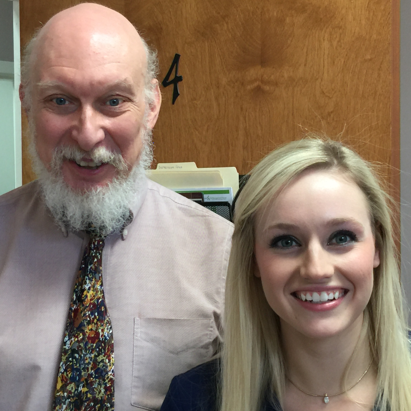 Portrait of a smiling elderly man with a white beard and a floral tie next to a smiling young blonde woman wearing a necklace.