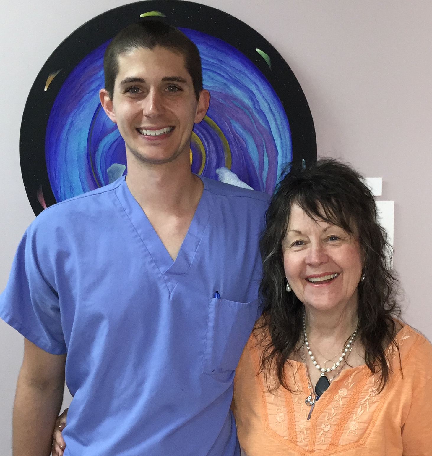 Smiling young man in blue scrubs standing next to smiling woman in an orange blouse with necklaces, in front of a circular blue and black artwork.