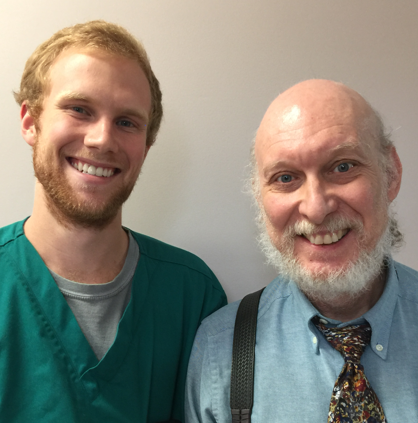 Smiling young man in green scrubs next to an older man with a white beard wearing a blue shirt and colorful tie.