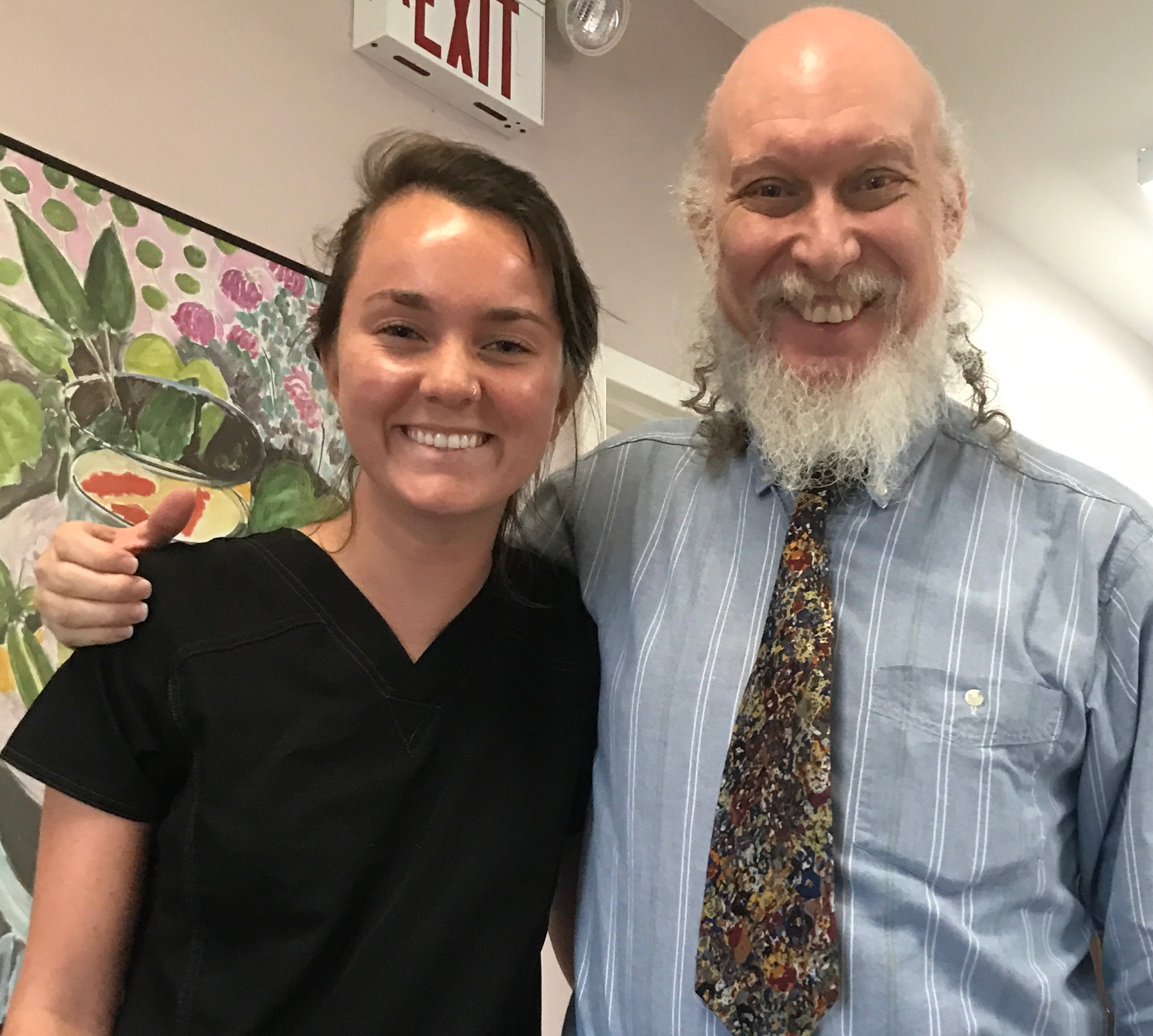 Smiling young woman in black scrubs and older man with beard wearing blue striped shirt and floral tie posing together indoors near a floral painting and exit sign.