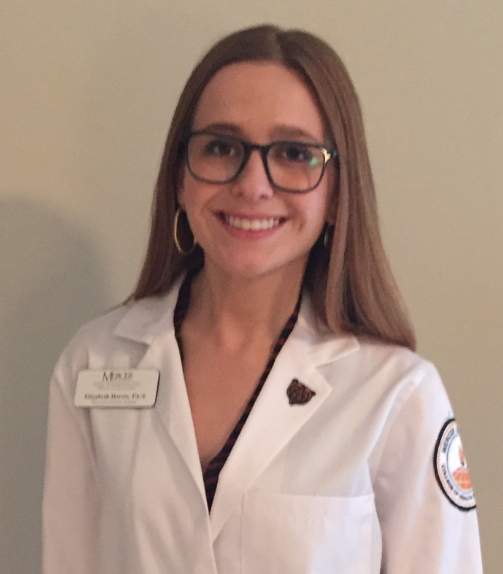 Smiling woman with long brown hair and glasses wearing a white lab coat with a name tag and embroidered patches.