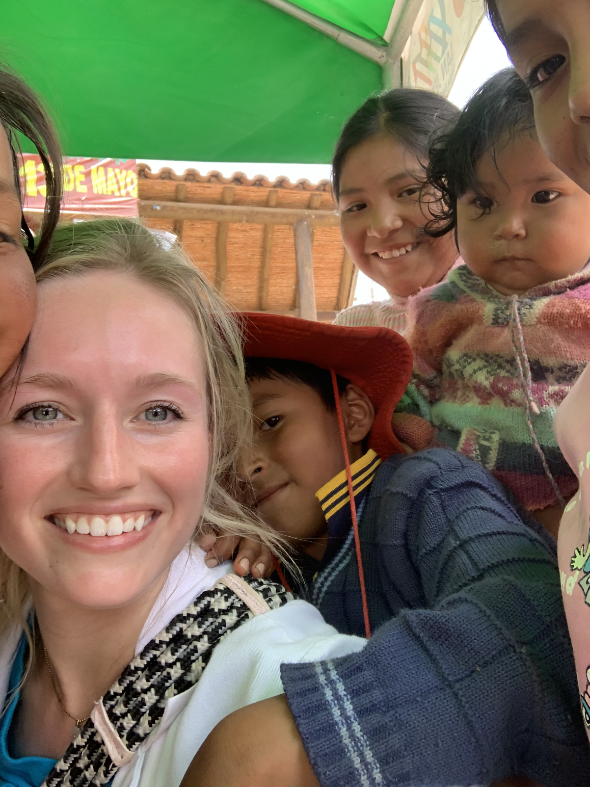 Group selfie of a smiling blonde woman with several children, including a boy in a red hat and a baby in a colorful sweater, under a green canopy.
